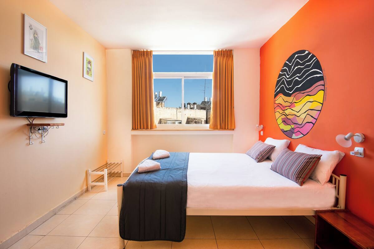 A bedroom with a bed, a television, a window with curtains, a wall art piece, and a view of buildings outside.