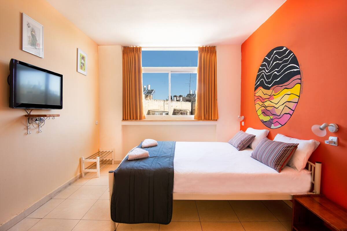 A bedroom with a bed, a television, a window with curtains, a wall art piece, and a view of buildings outside.