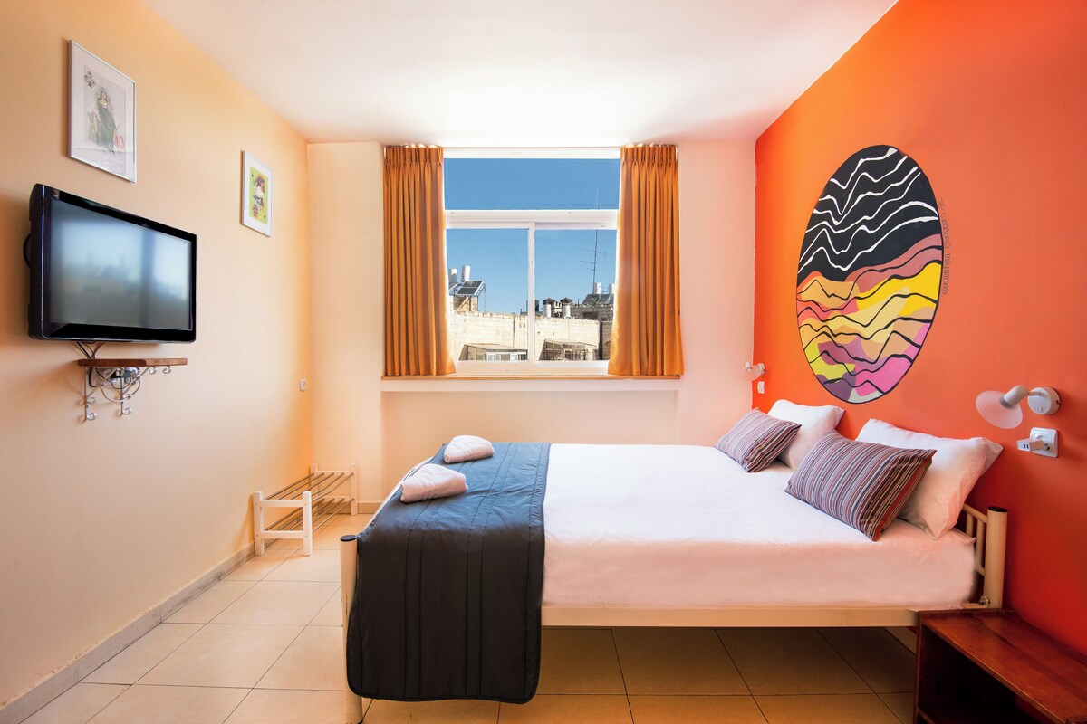 Un dormitorio con cama, televisor, ventana con cortinas, una obra de arte en la pared y vistas a edificios.