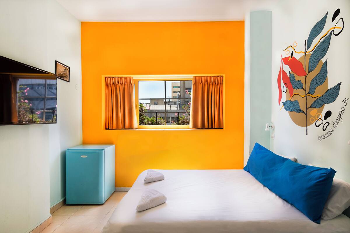 A bedroom with a bed, a small blue refrigerator, a window with curtains, and a wall art piece.