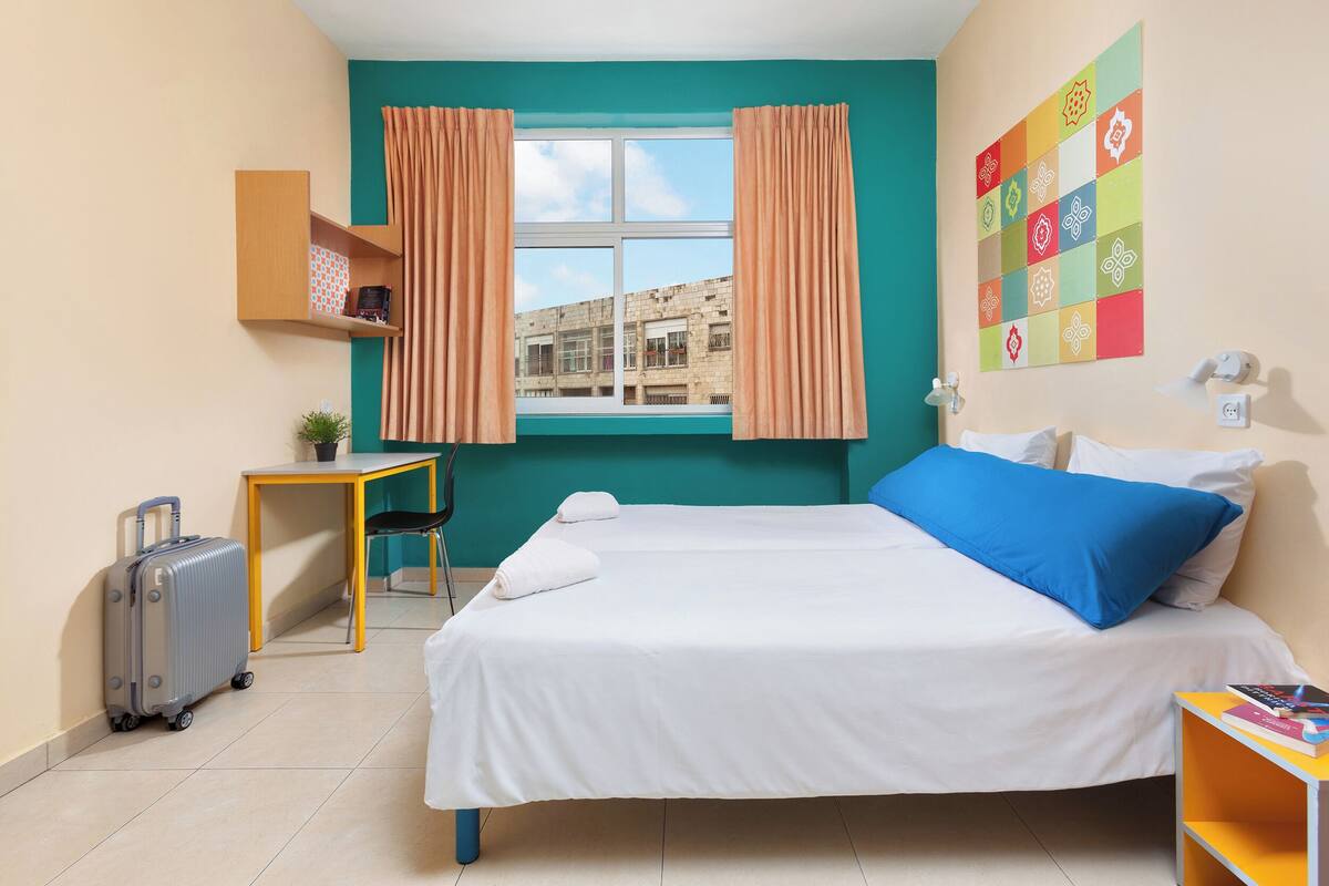 A hotel room with a bed, a desk, a suitcase, a window with curtains, and a wall with a colorful pattern.
