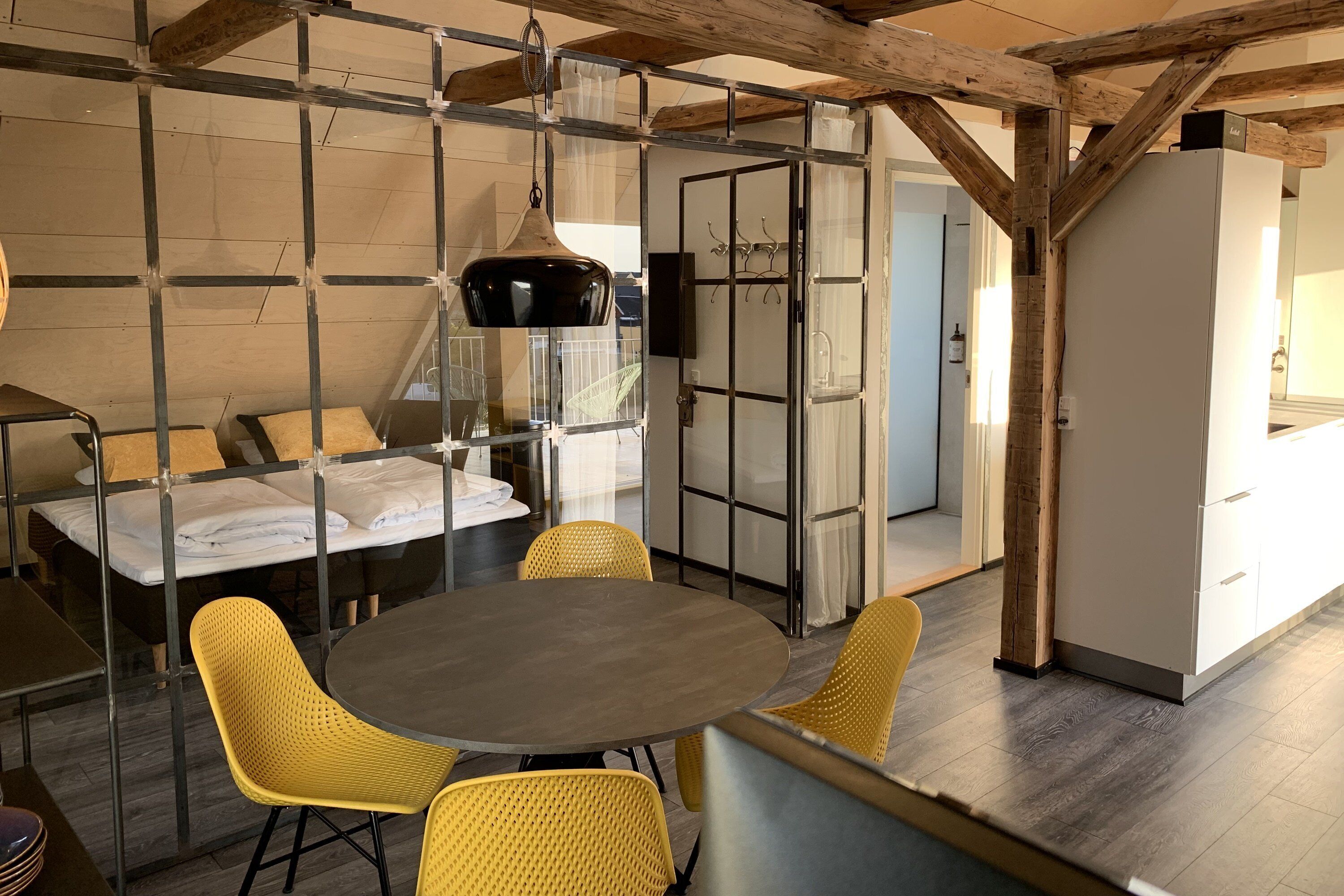 Een kamer met een ronde tafel, gele stoelen, een bed met witte lakens en een moderne keukenhoek.