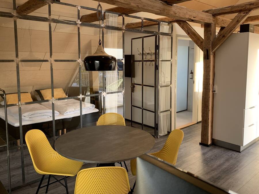 Een kamer met een ronde tafel, gele stoelen, een bed met witte lakens en een moderne keukenhoek.
