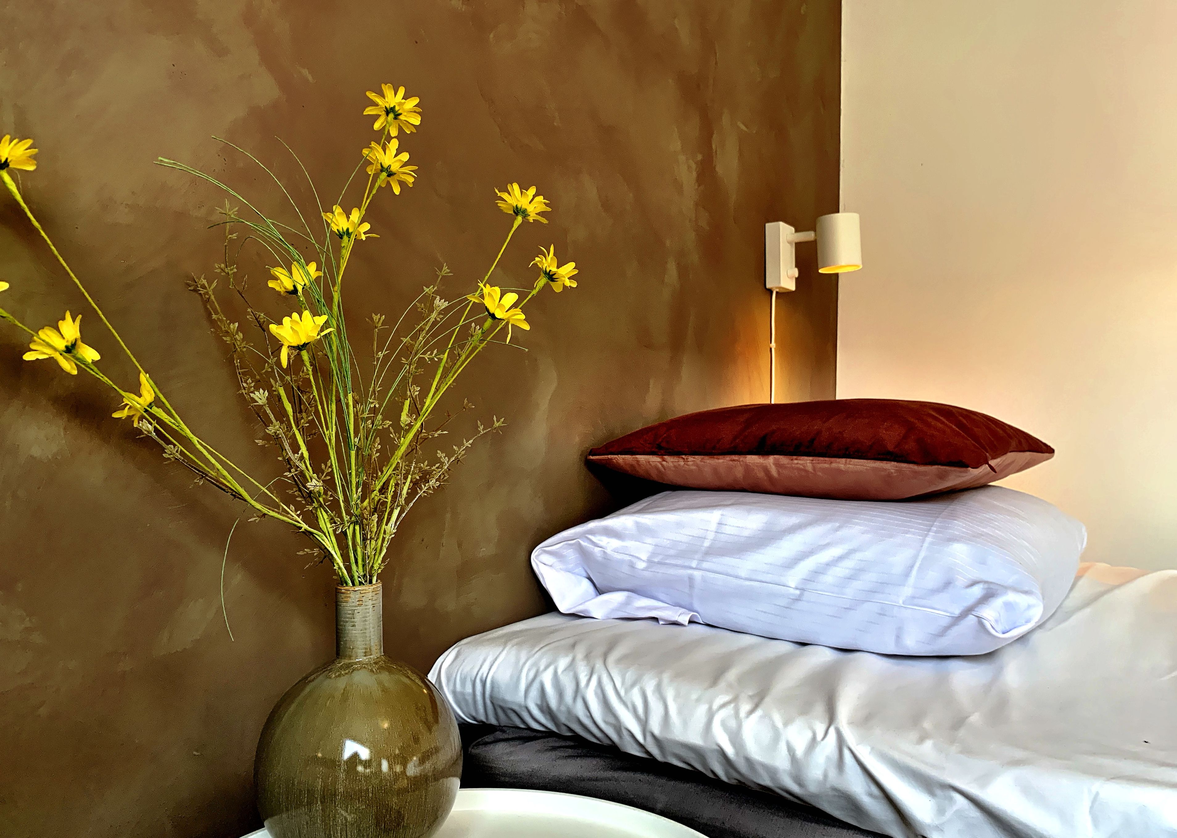 A bed with white bedding, a red pillow, and a vase with yellow flowers on a bedside table.