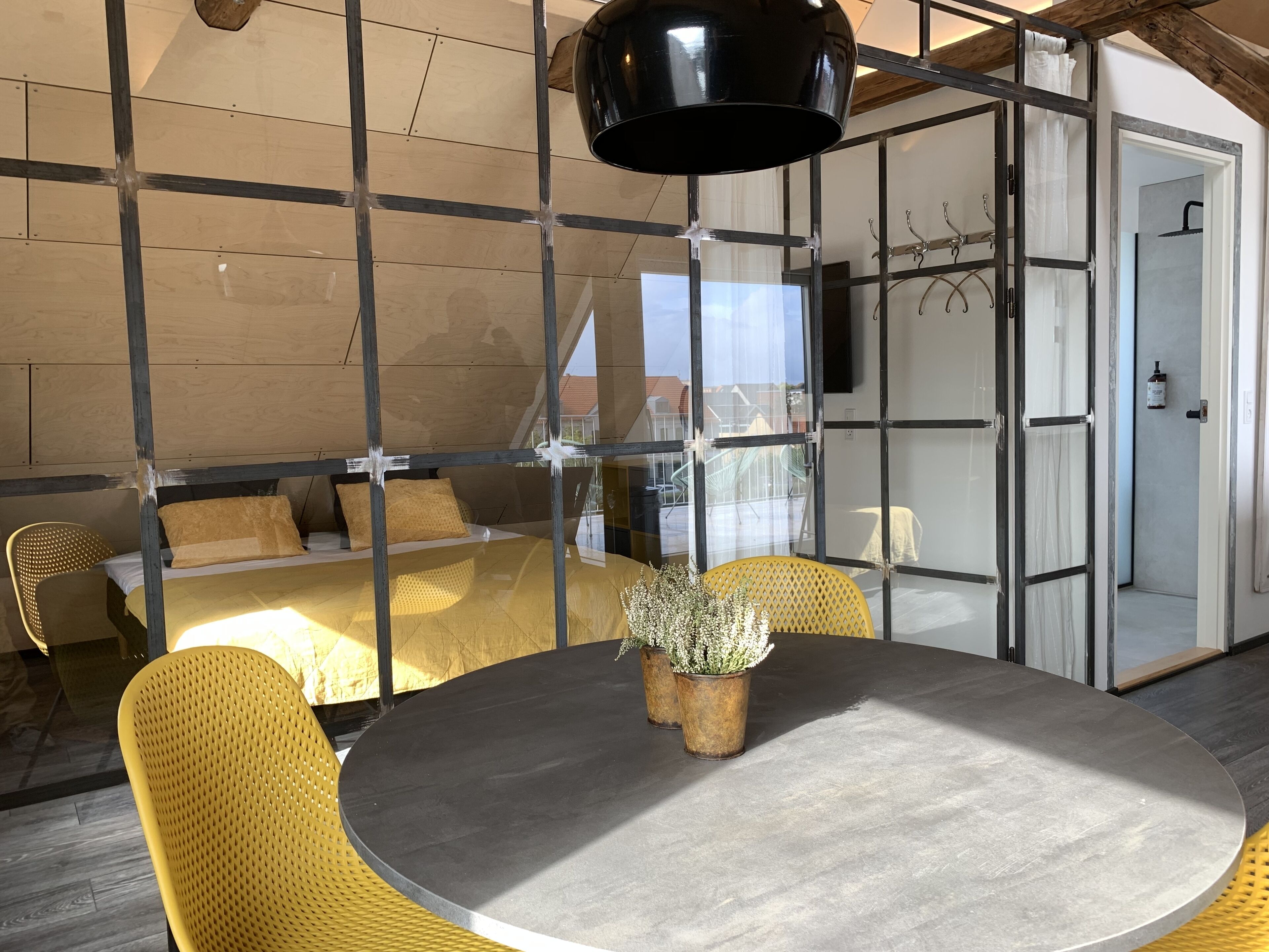 A modern room with a round table, yellow chairs, a bed with a wooden headboard, and a large window.