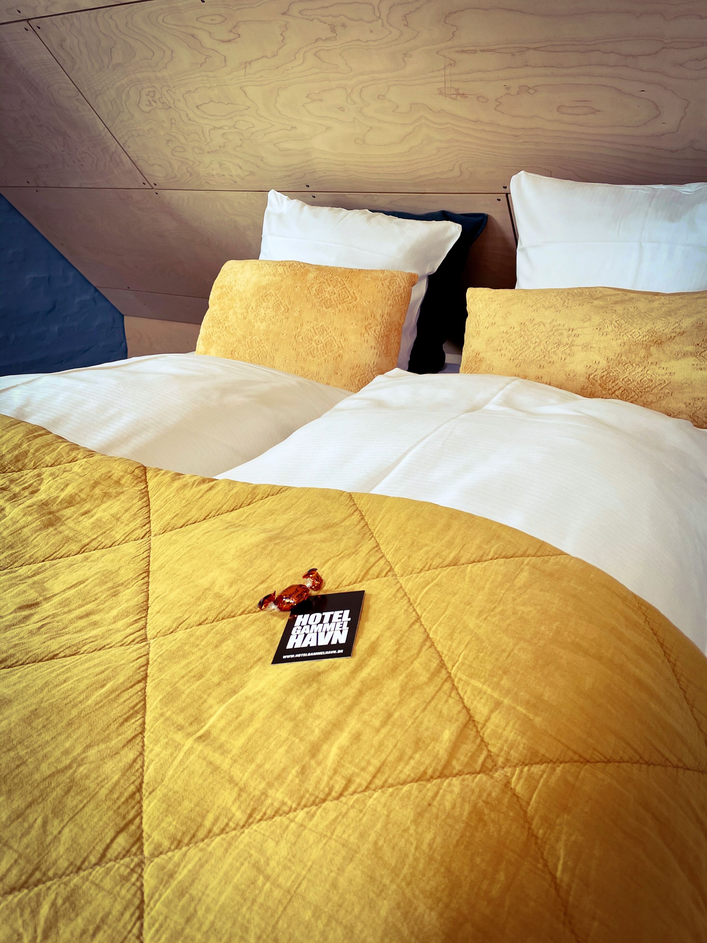 A hotel room with a bed featuring a yellow quilted bedspread, white pillows, and a black tag on the bed.