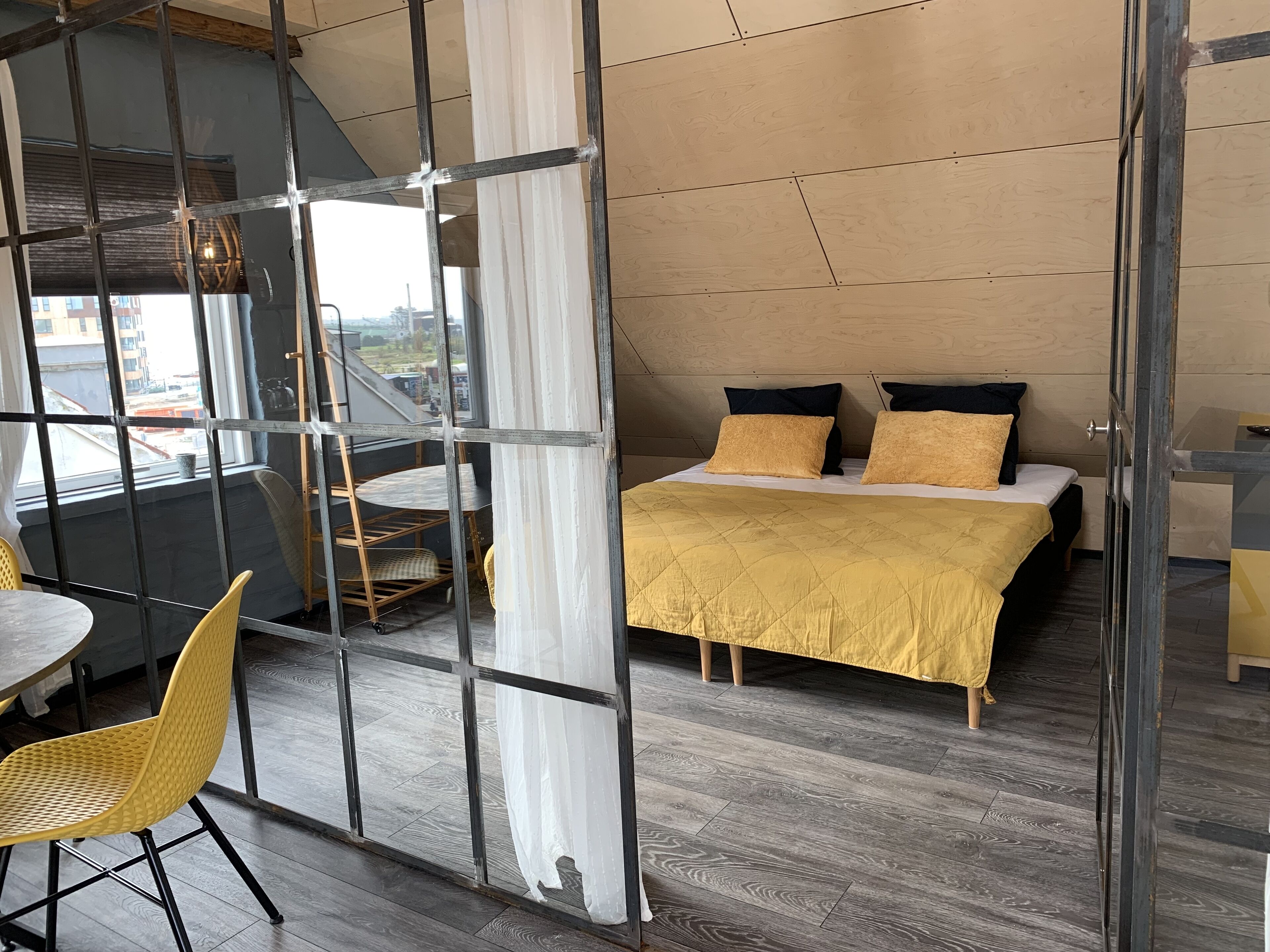 A modern bedroom with a bed, two pillows, a yellow blanket, a wooden floor, and a view through a glass partition.