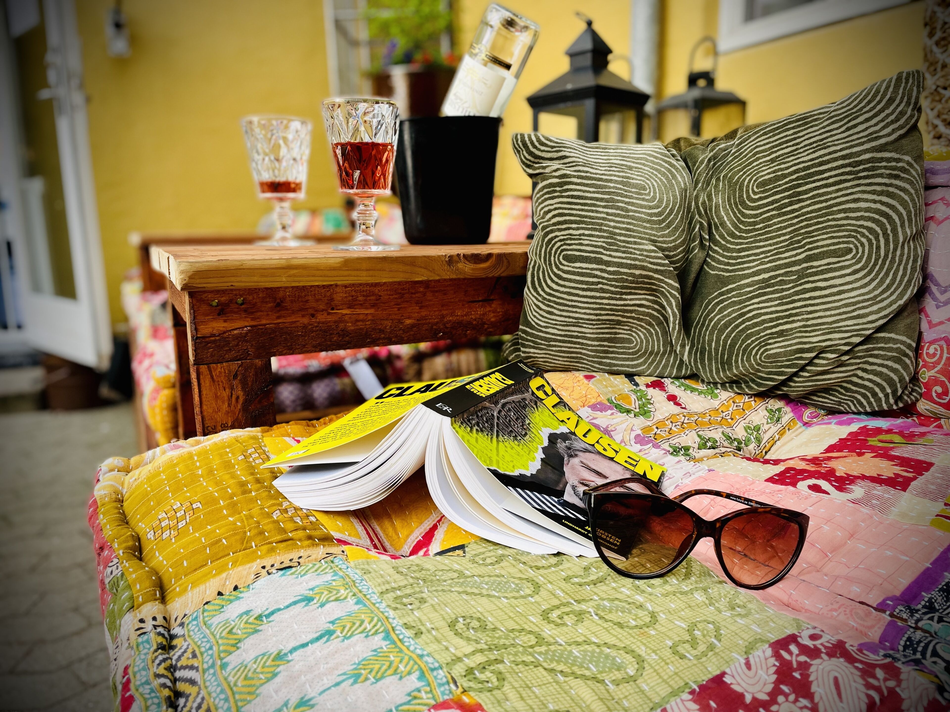 A bed with a quilt, a magazine, a pair of sunglasses, and a bottle of perfume on a wooden nightstand.