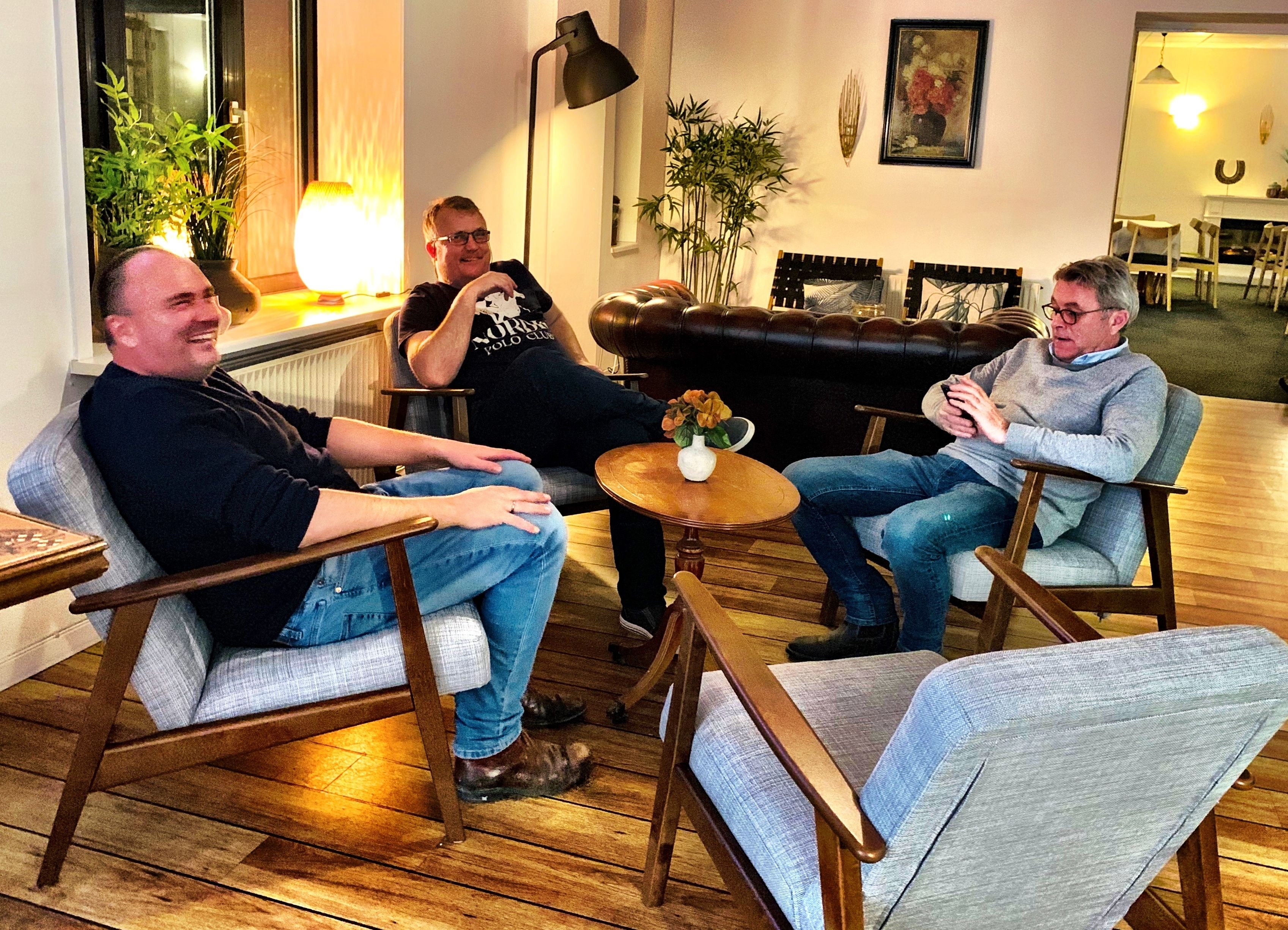 Eine Gruppe von Personen sitzt in einem hellen Raum mit Holzboden, Ledersofas und einem kleinen Tisch mit Blumenvase.