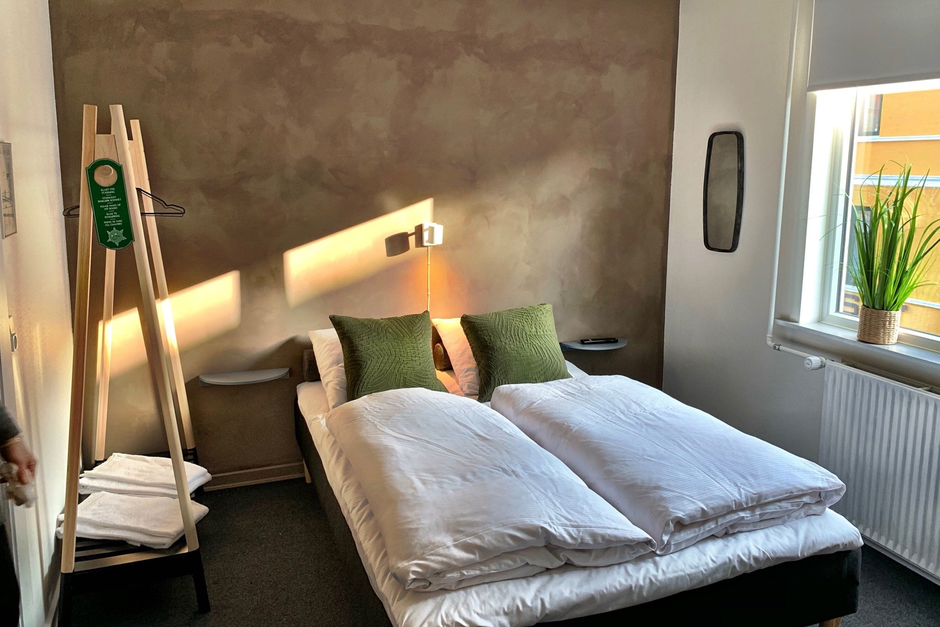 A neatly made bed with white linens, a bedside table with a lamp, and a window with a plant.