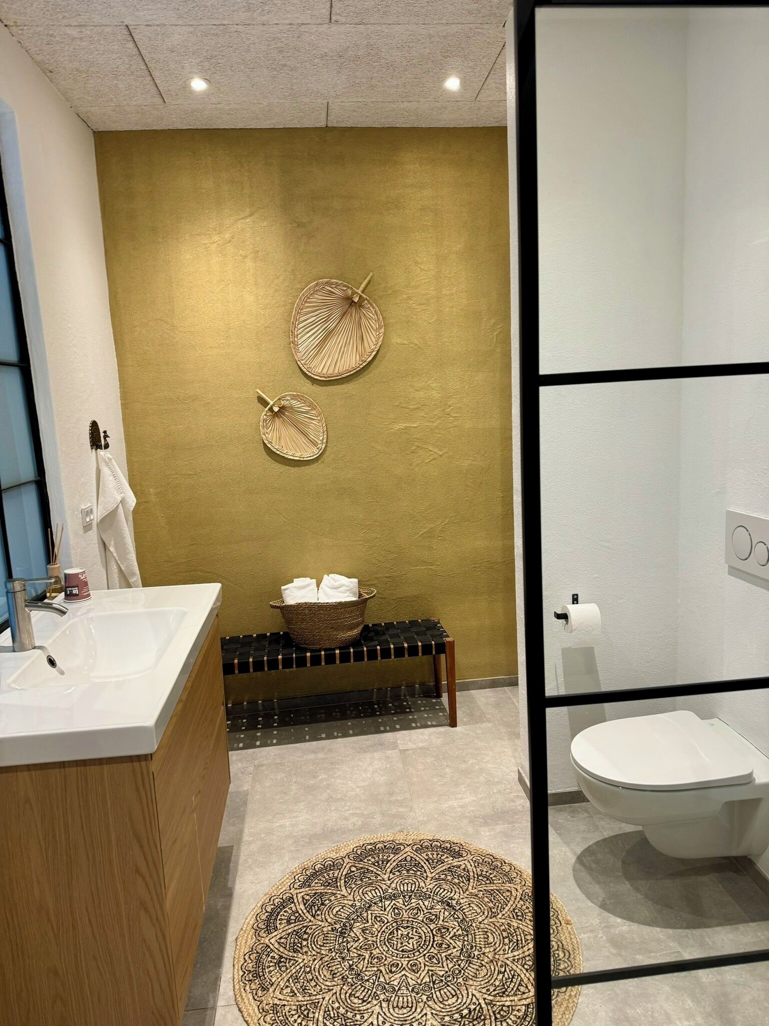 Une salle de bain moderne avec un mur d’accent doré, un meuble-lavabo en bois, un miroir rond et un banc à carreaux noirs et blancs.