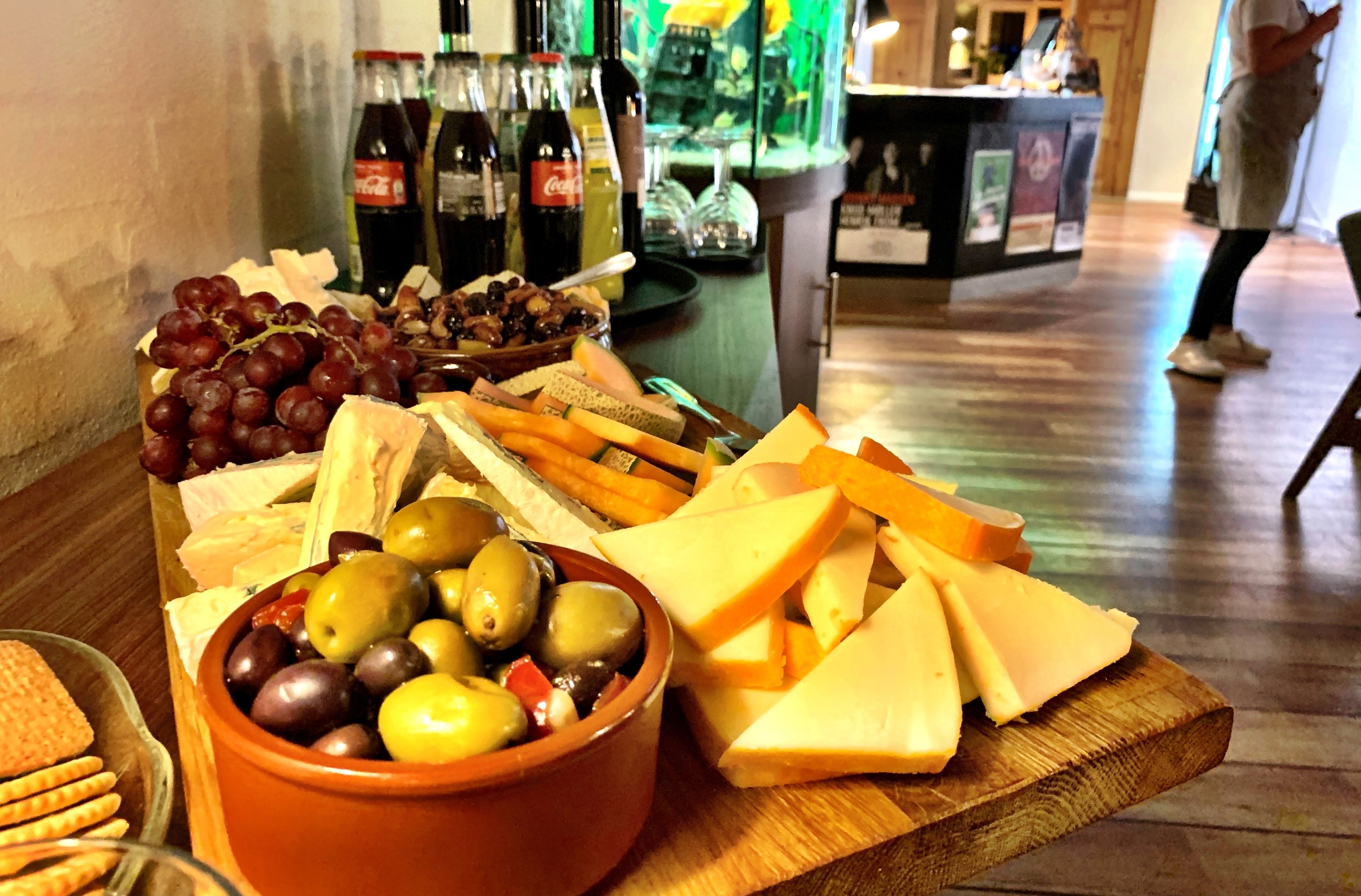 Una tabla de madera con una fuente de queso y fruta, y botellas de vino al fondo.