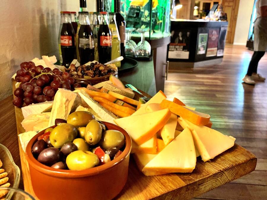 Una tabla de madera con una fuente de queso y fruta, y botellas de vino al fondo.