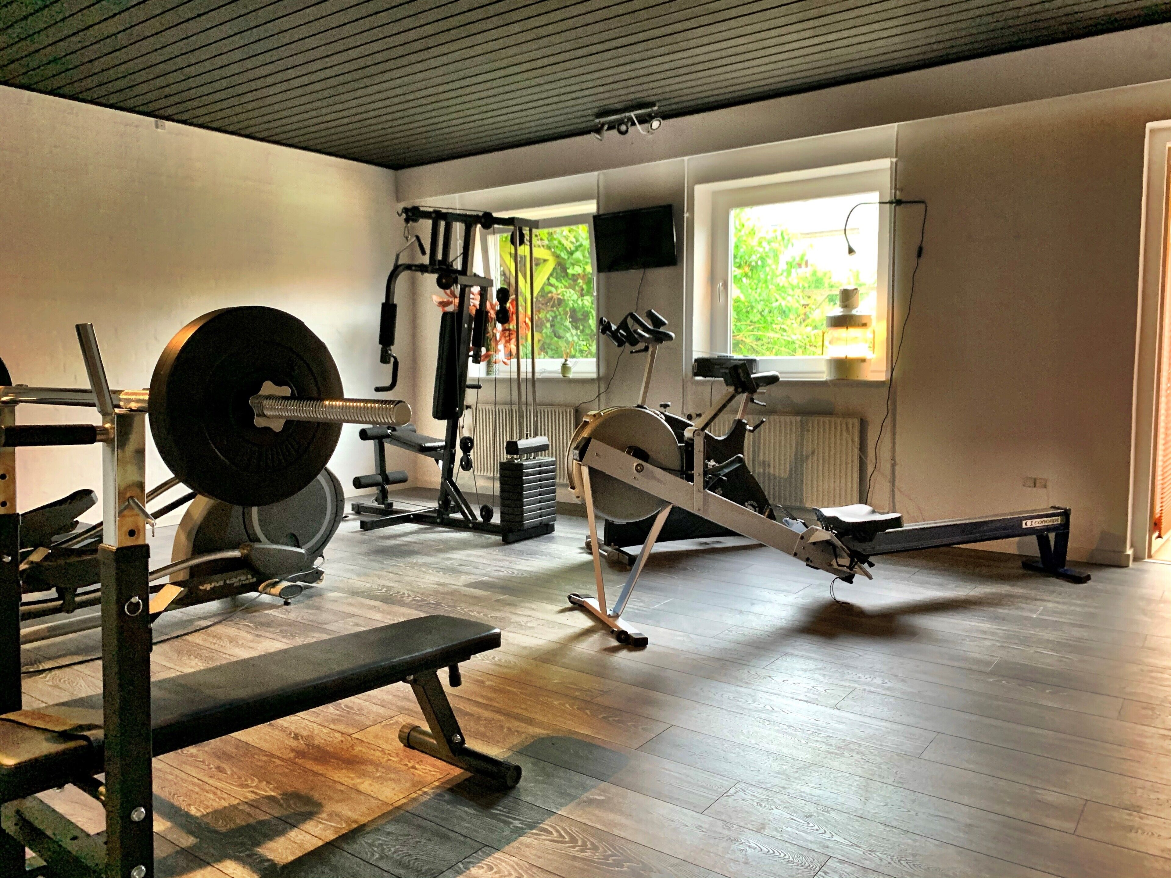 Un gimnasio con banco, estante para pesas, máquina de remo y televisor en la pared.