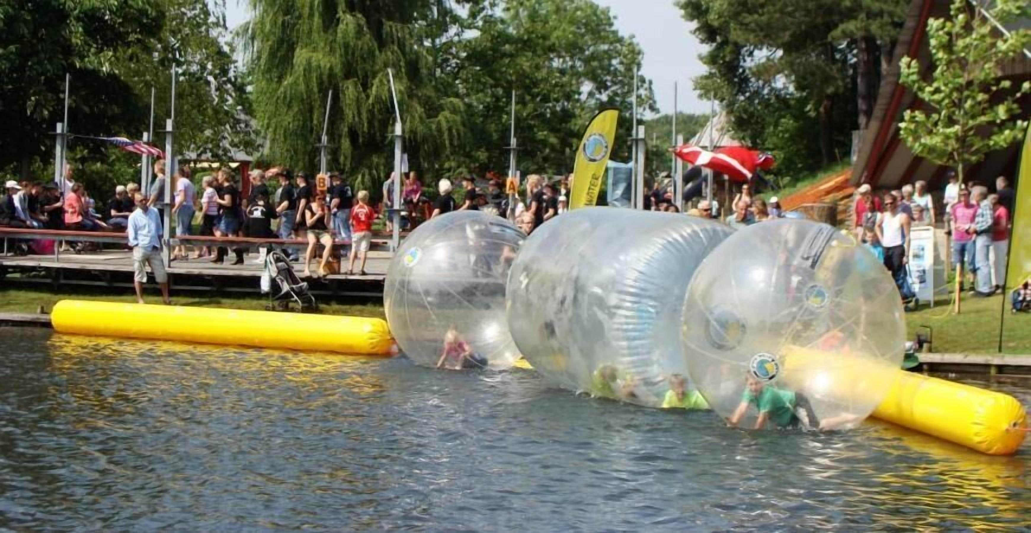 Personas en esferas transparentes sobre el agua, espectadores en un muelle y una barrera inflable amarilla.