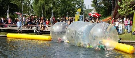 Personas en esferas transparentes sobre el agua, espectadores en un muelle y una barrera inflable amarilla.
