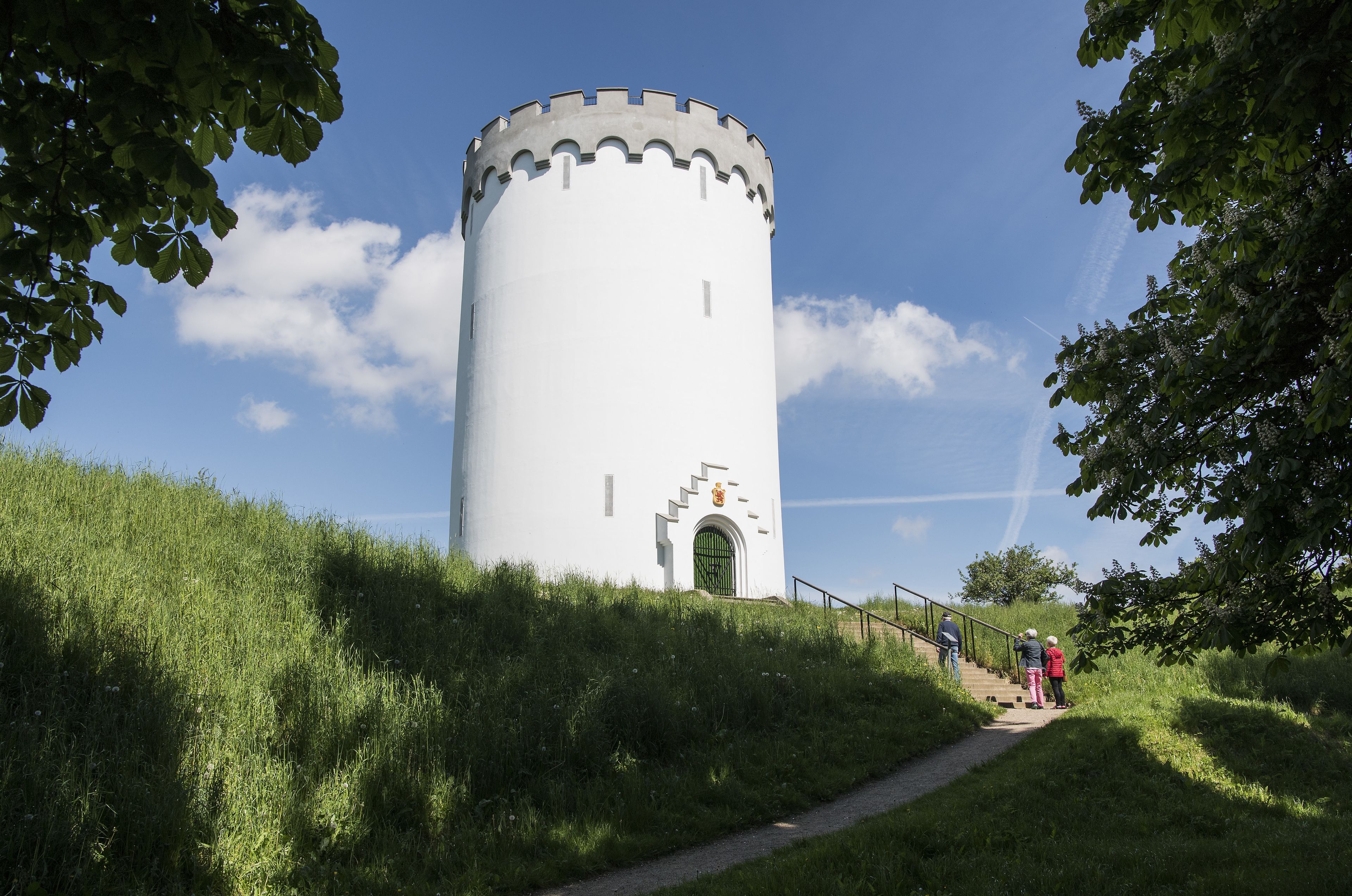 A white tower with a crenellated top, surrounded by greenery and a clear sky.