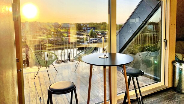 A modern room with a large sliding glass door leading to a balcony with a view of a cityscape and a river. There is a round table with two stools and a candle on it. - Hotel Gammel Havn - Good Night Sleep Tight (Fredericia)