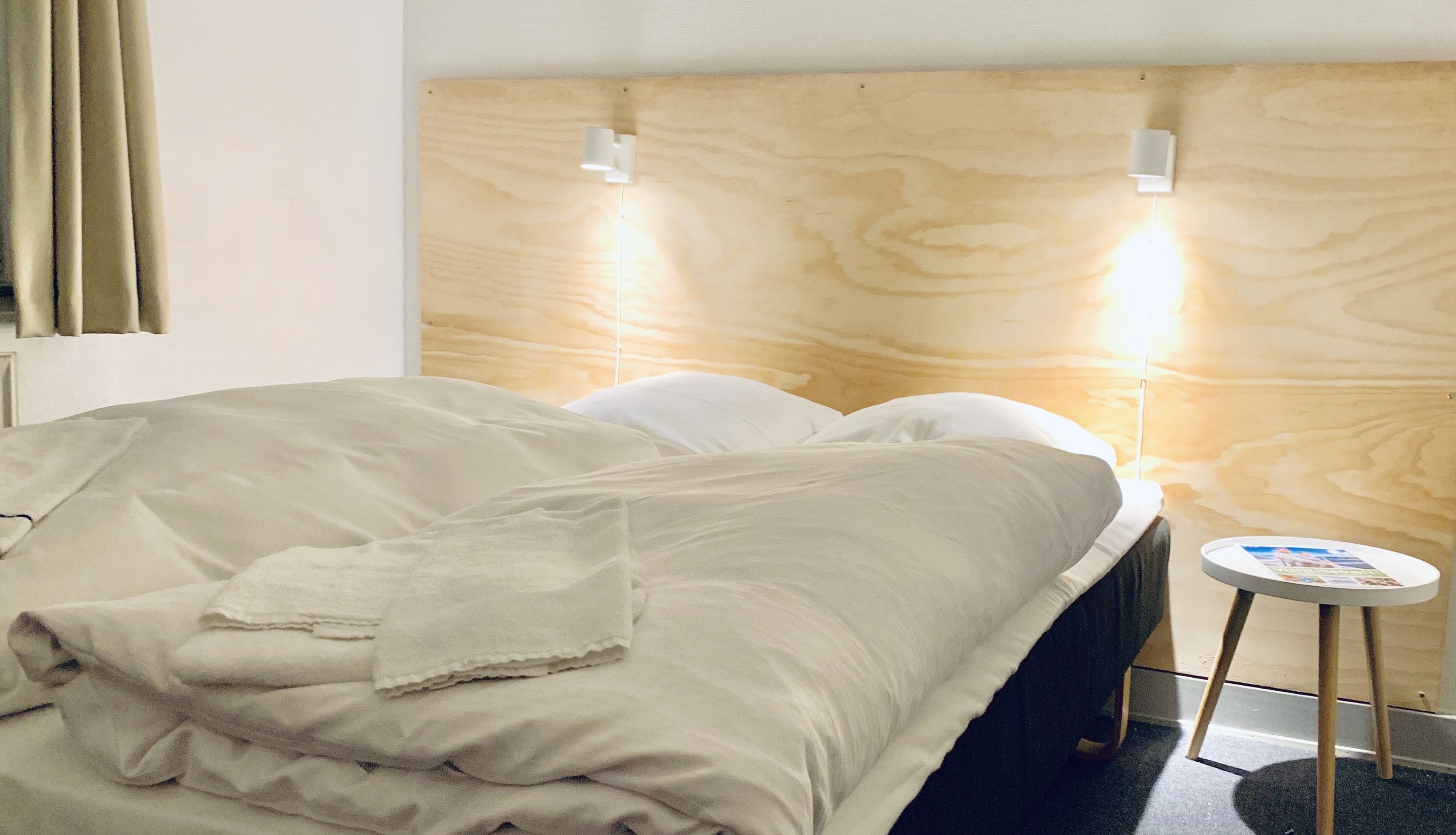 A neatly made bed with a white comforter and pillows, a small round table with a magazine, and a wooden headboard with wall-mounted lights.