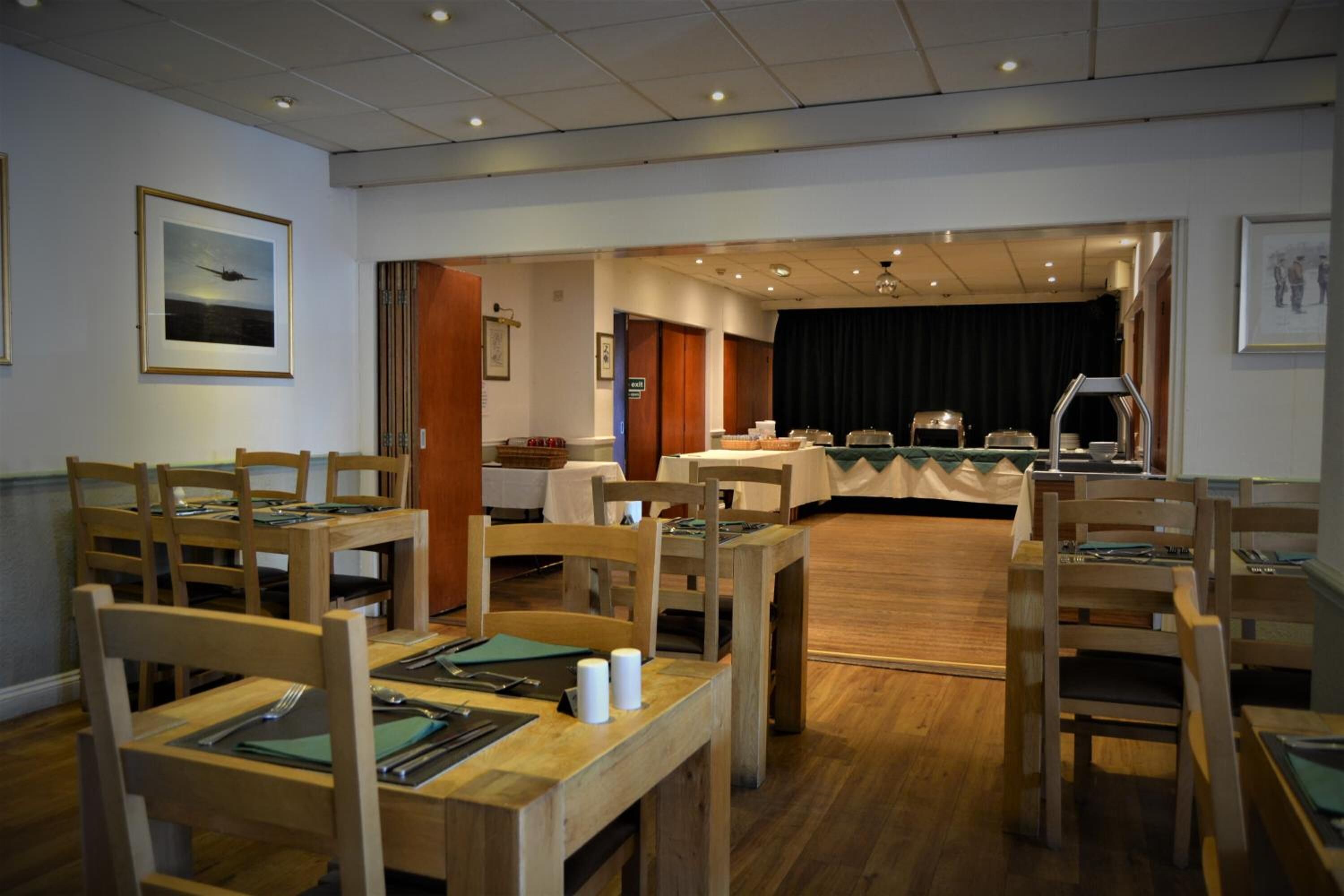 A hotel dining area with wooden tables and chairs, a stage with a black backdrop, and framed artwork on the walls.
