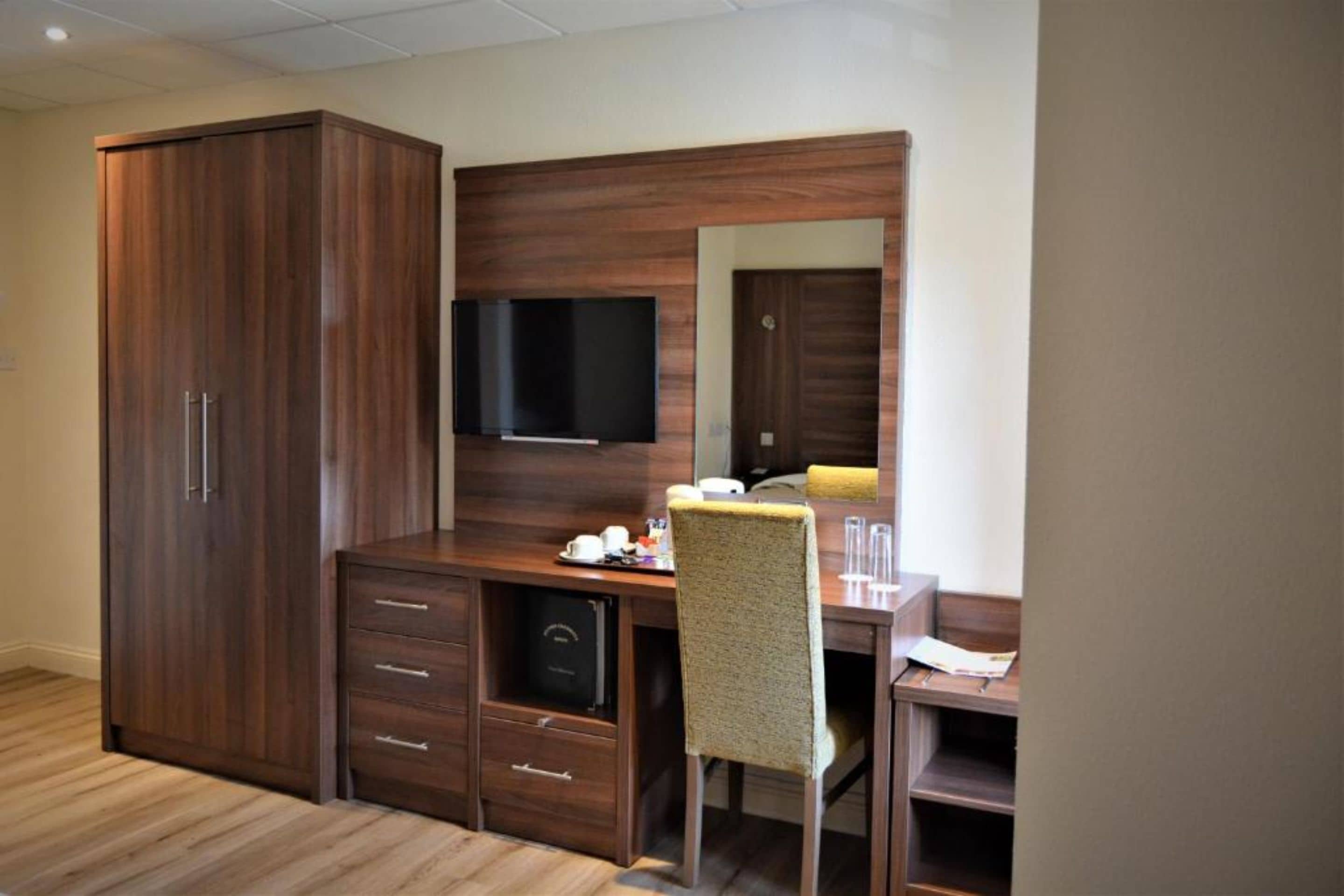 Quarto de hotel com guarda-roupa de madeira, uma escrivaninha com cadeira, televisão de tela plana e um espelho.