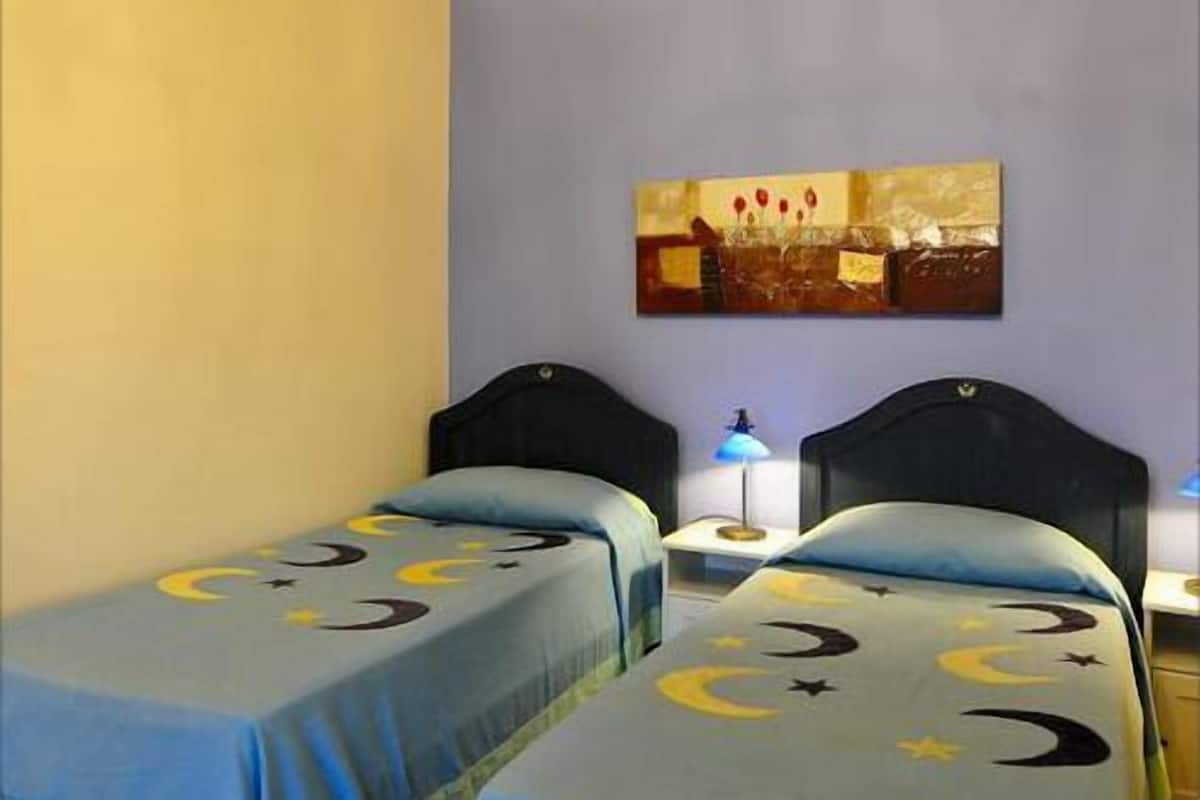 Une chambre avec deux lits simples, chacun recouvert d’une couverture bleue ornée de motifs représentant la lune et les étoiles. Un tableau représentant un paysage avec des fleurs rouges est accroché au mur.