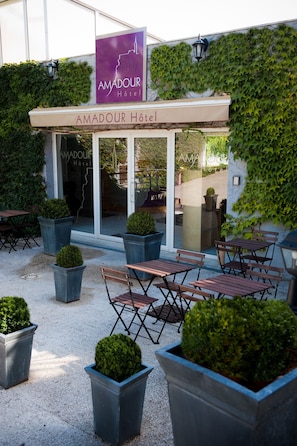 An outdoor seating area with tables and chairs, surrounded by planters with shrubs. - Amadour Hôtel (Rocamadour)