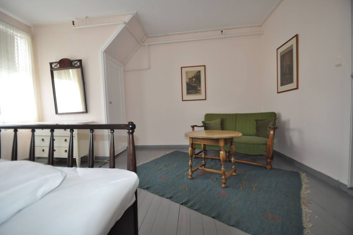 A bedroom with a bed, a green armchair, a wooden table, and two framed pictures on the wall.
