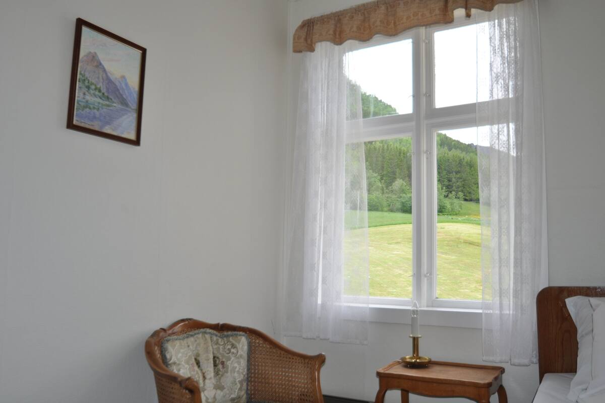A room with a large window, a framed painting, a wicker chair, a bedside table with a lamp, and a bed.
