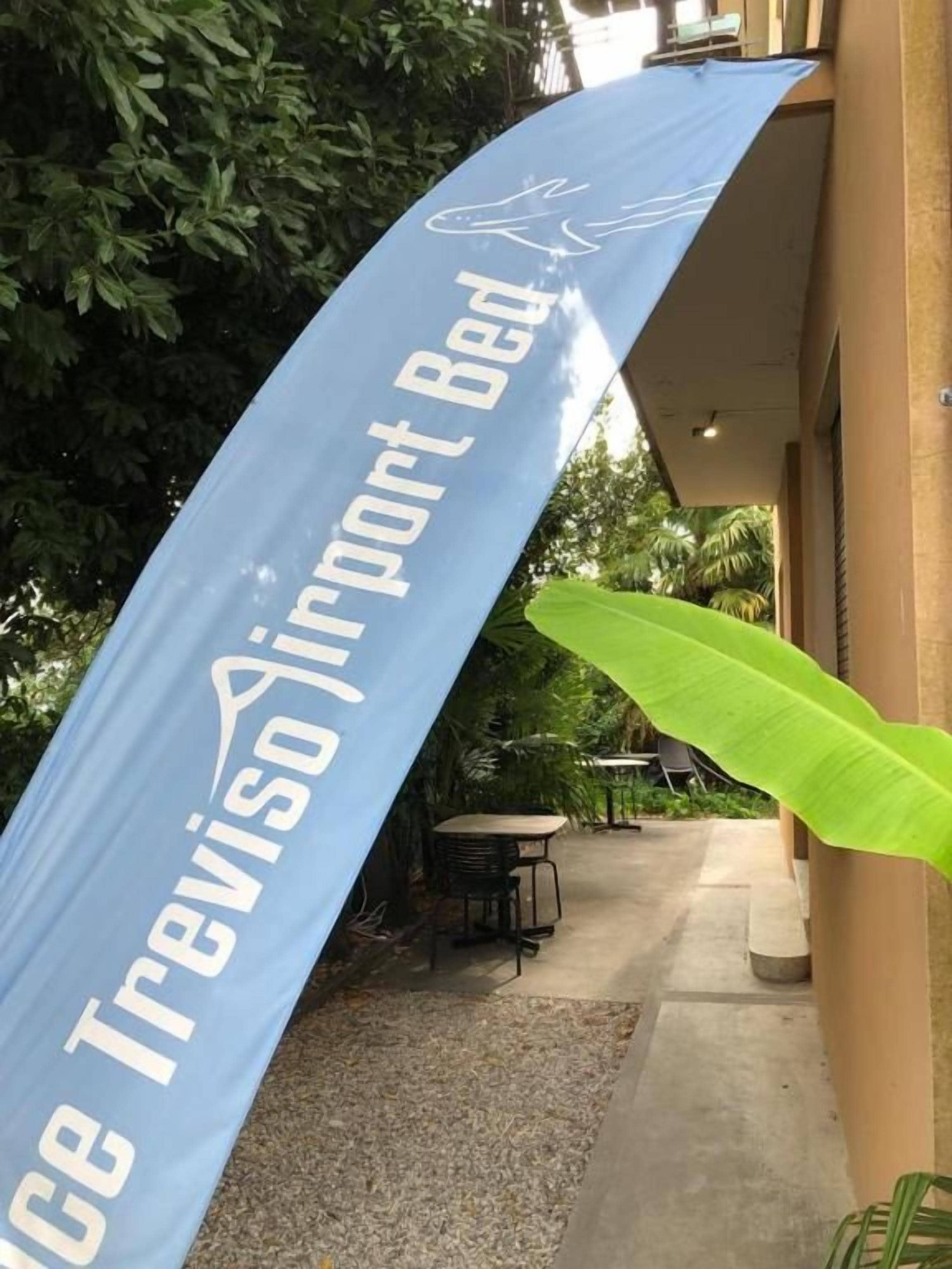 A blue banner with 'Treviso Airport Bed' text, a concrete pathway, a table, chairs, and a large green plant.