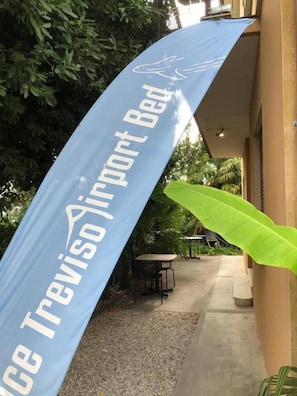 Un banner azul con el texto 'Treviso Airport Bed', un camino de hormigón, una mesa, sillas y una planta verde grande.