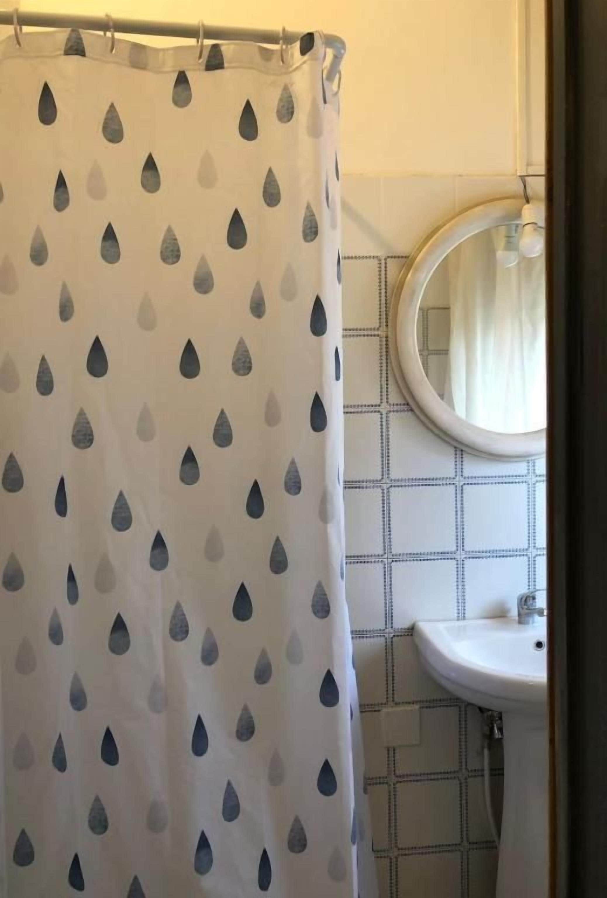 Ein Badezimmer mit Duschvorhang mit Tropfenmuster, einem runden Spiegel und einem Waschbecken mit gefliestem Rücksprung.