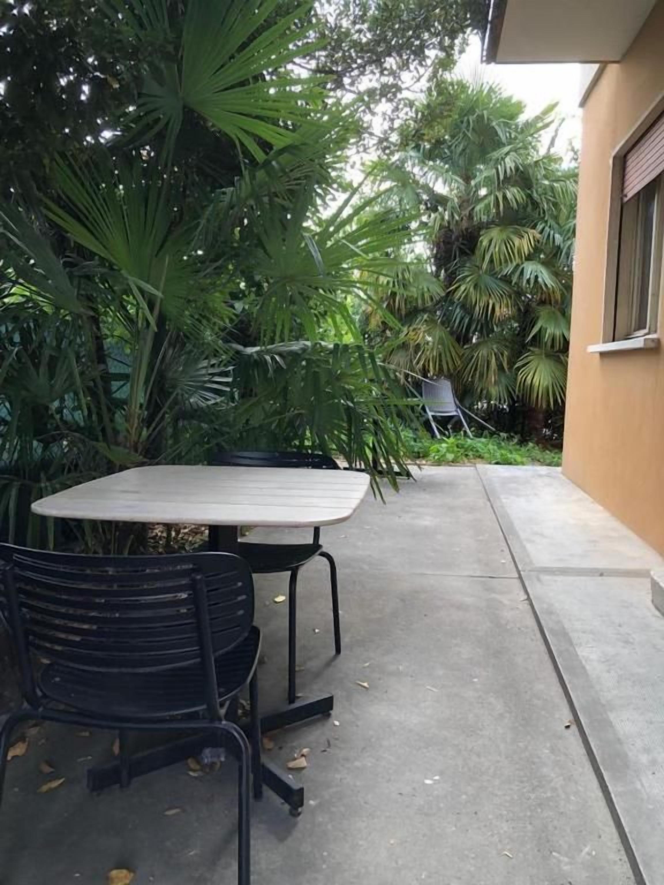 An outdoor patio with a table and chairs, surrounded by lush greenery and a building.