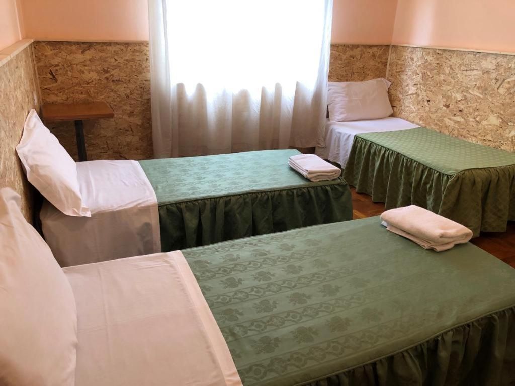 Ein Zimmer mit zwei Einzelbetten, jeweils mit einer grünen Tagesdecke und weißen Kissen. Ein kleiner Tisch und ein Fenster mit Vorhängen sind ebenfalls vorhanden.