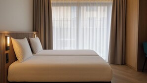 Une chambre d’hôtel dotée d’un grand lit, d’une tête de lit avec éclairage intégré et offrant une vue sur la ville à travers des rideaux transparents.