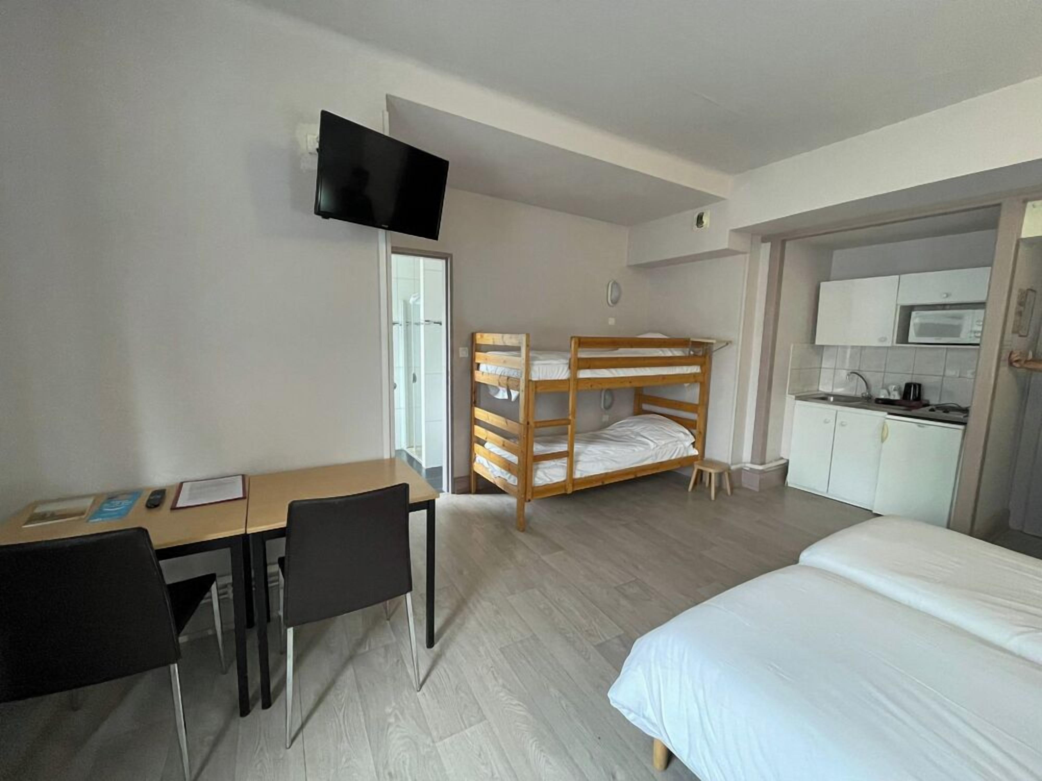 Une chambre avec un lit superposé, un bureau avec des chaises, un téléviseur fixé au mur et un coin cuisine.