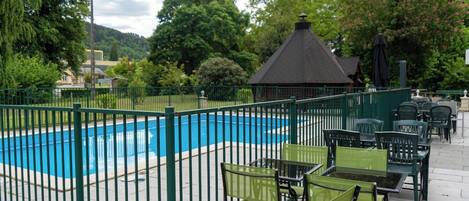 A swimming pool surrounded by a green fence, with outdoor seating and a gazebo in the background.