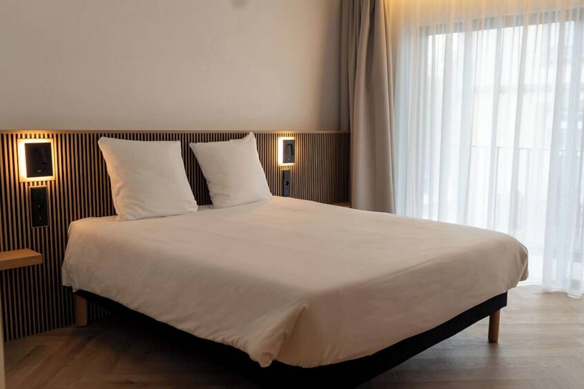 A modern hotel room with a large bed, two bedside tables with lamps, and a large window with sheer curtains.