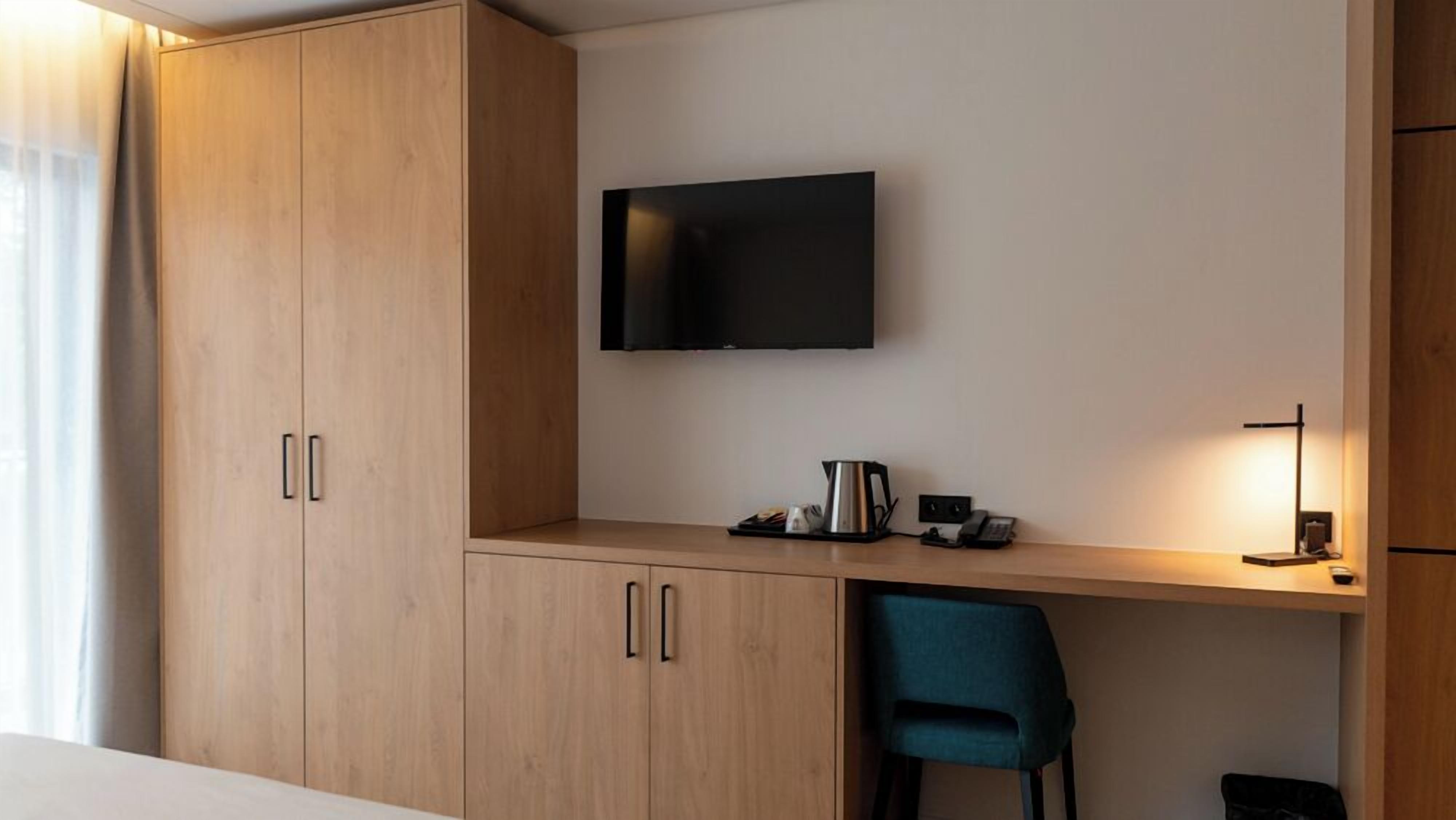 A hotel room with a wooden wardrobe, a desk with a flat-screen TV, a chair, and a bedside table with a lamp.