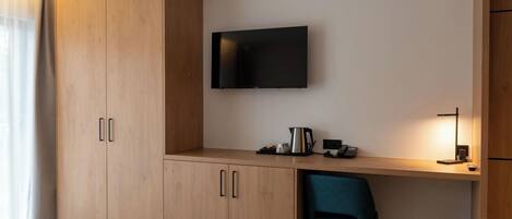 A hotel room with a wooden wardrobe, a desk with a flat-screen TV, a chair, and a bedside table with a lamp.