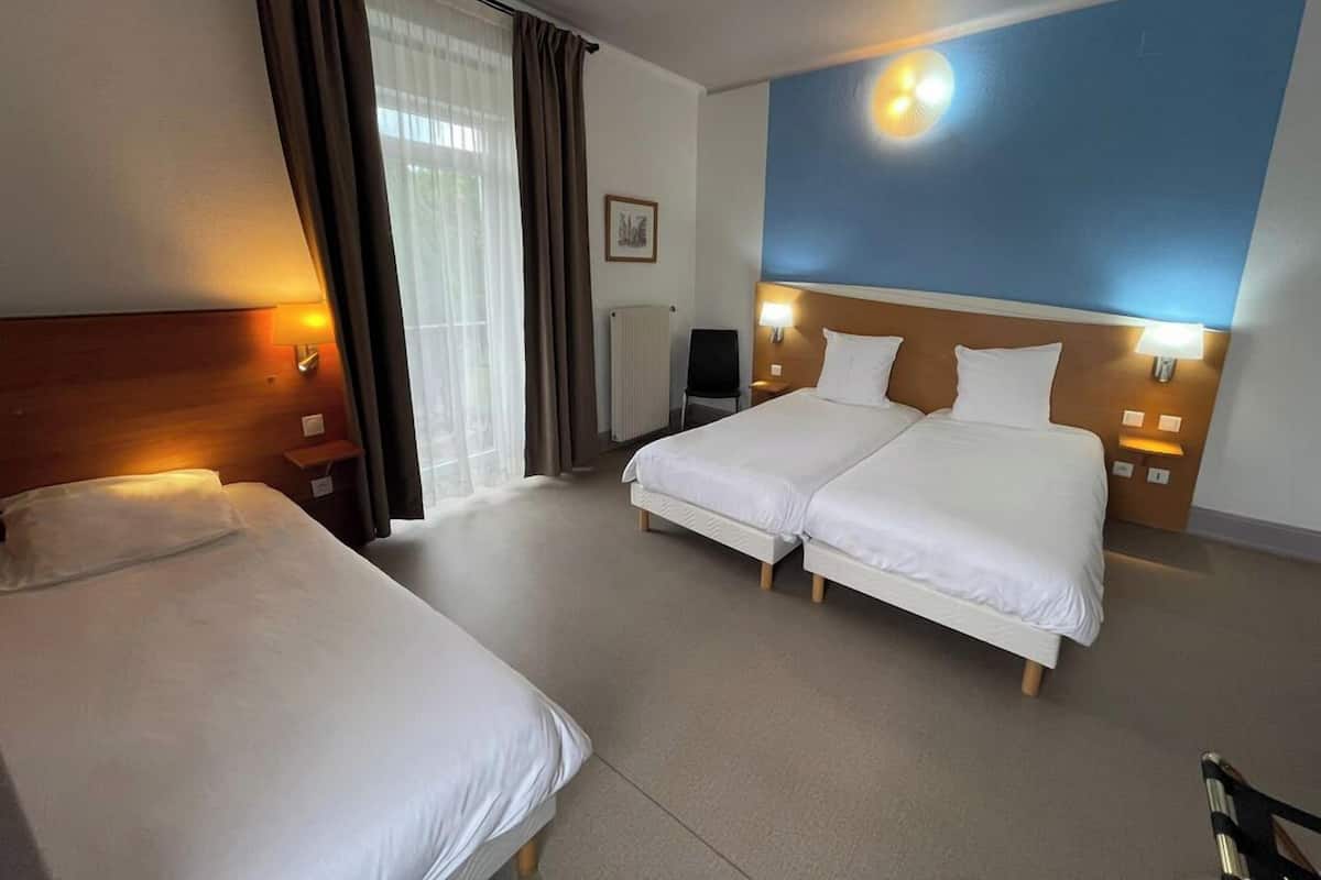 A hotel room with two single beds, a double bed, a desk, a chair, and a large window with curtains.