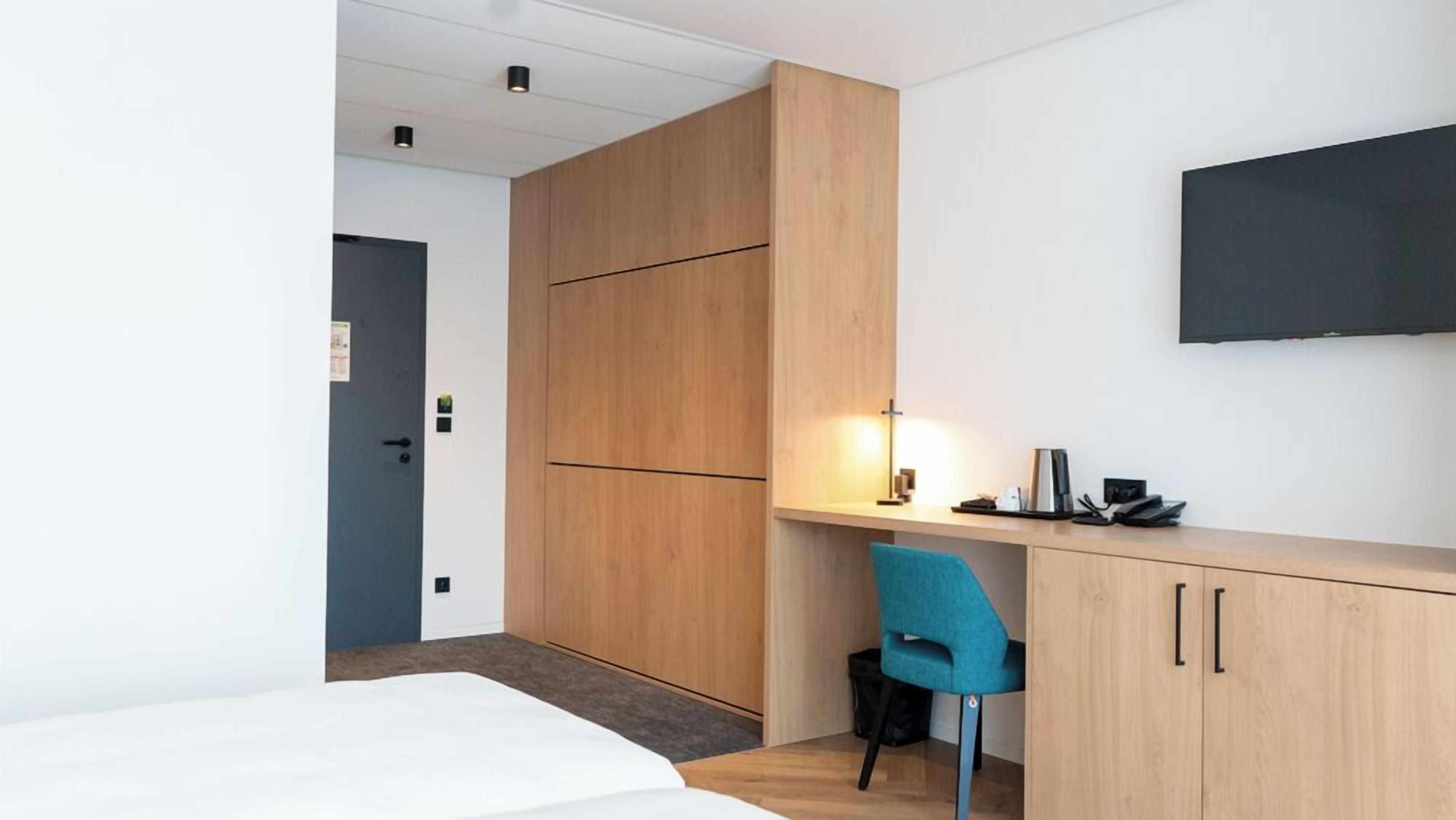 A modern hotel room with a bed, a desk with a chair, a flat-screen TV, and a door.