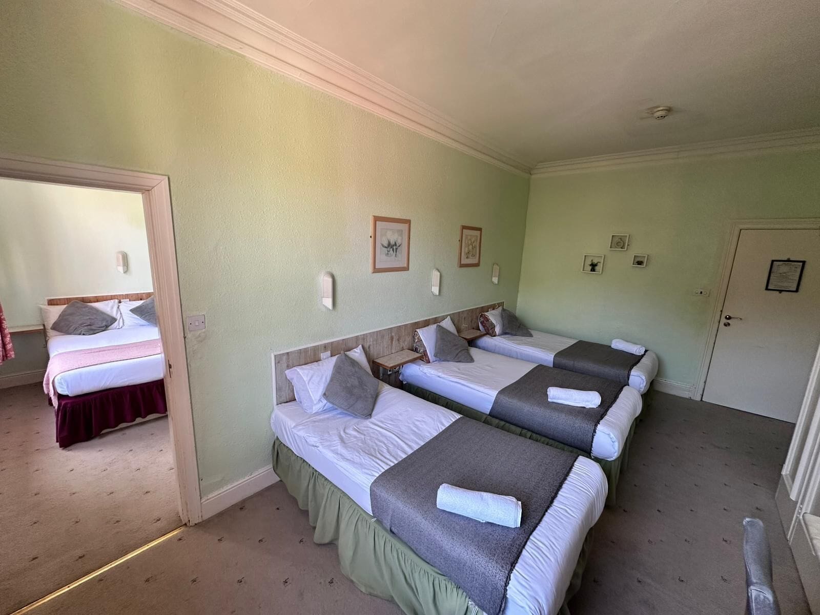 A hotel room with three single beds, a door, a mirror, and a small table.