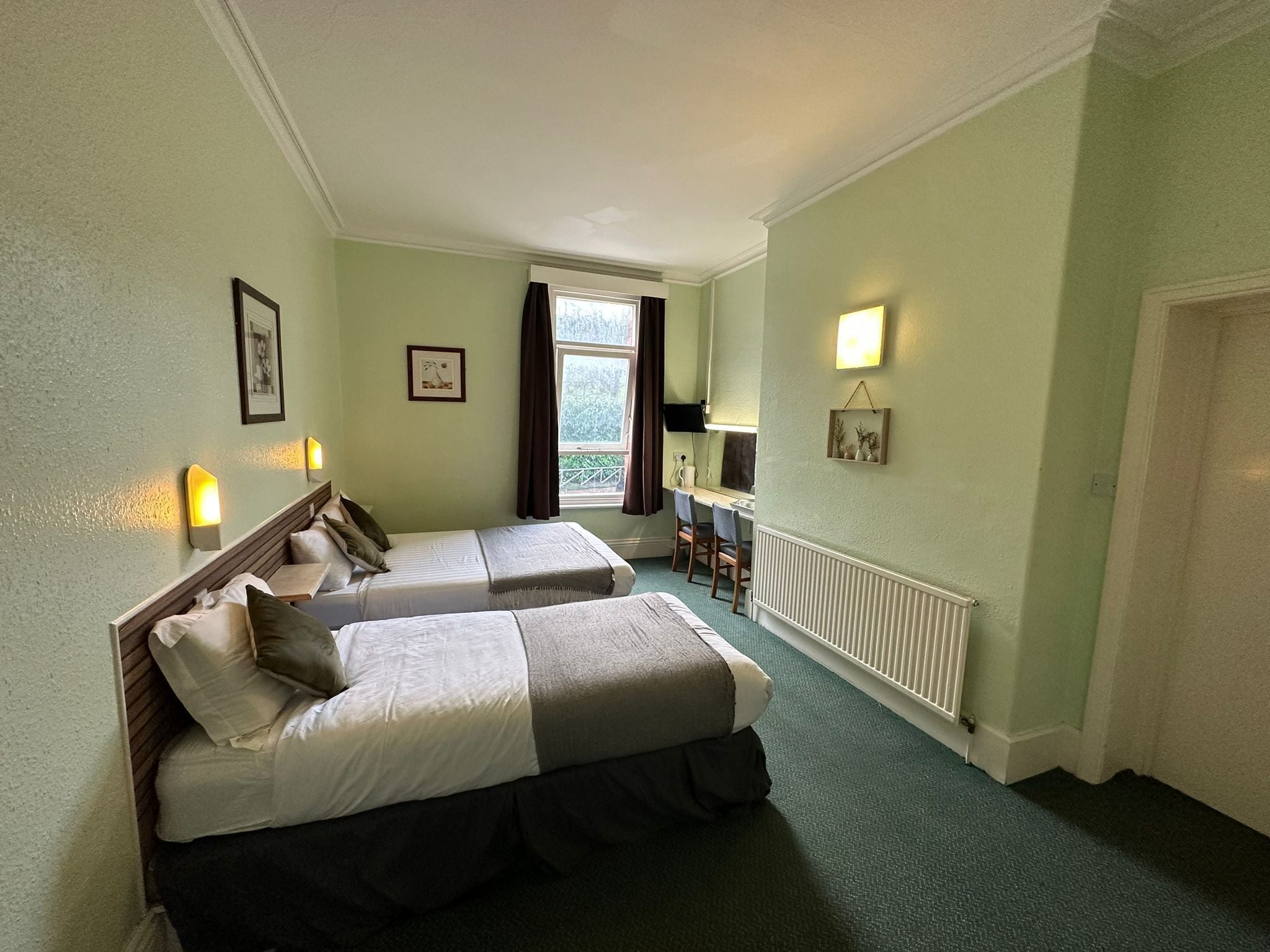 A hotel room with two beds, a desk, a chair, a window with curtains, and a radiator.