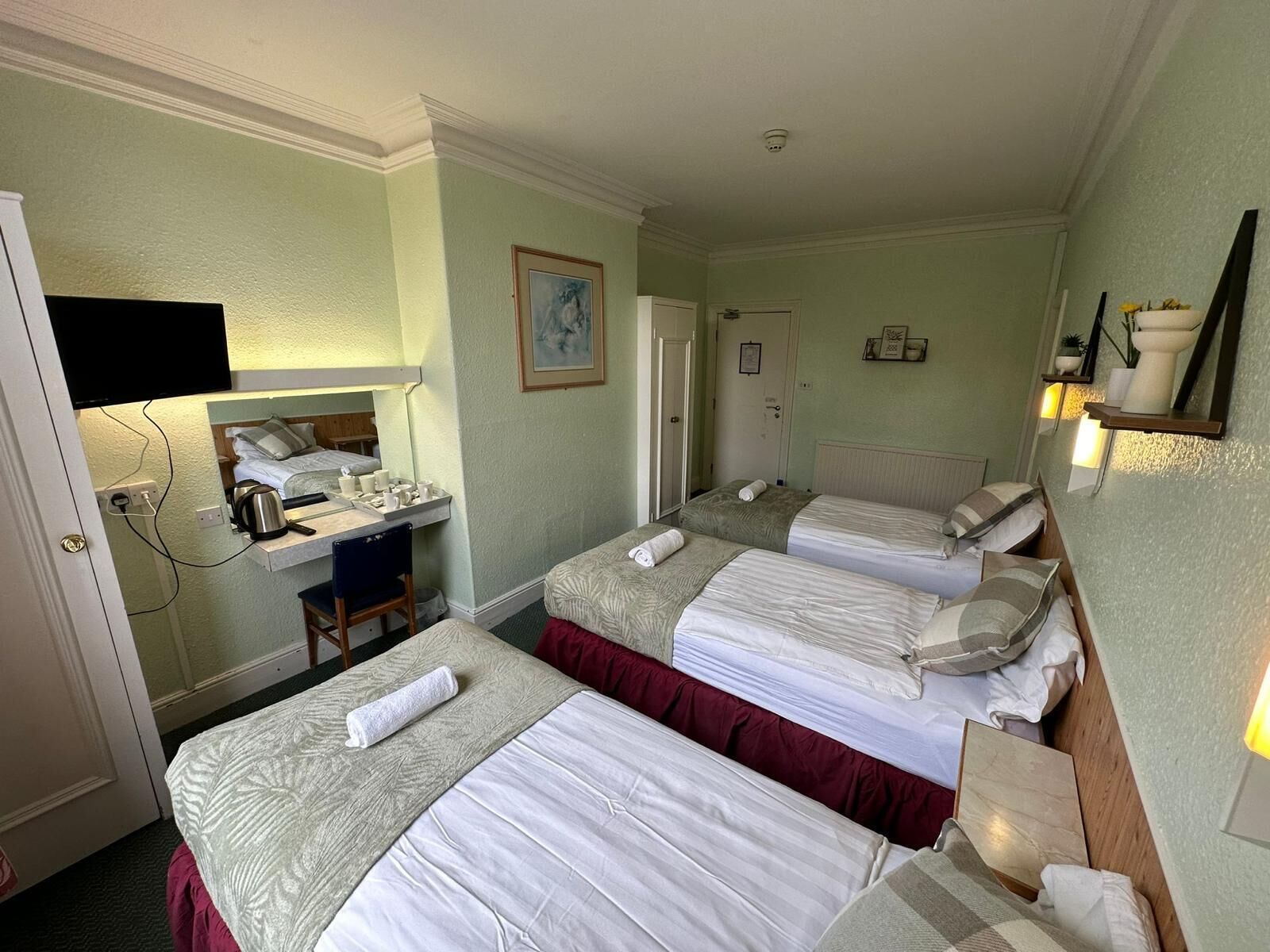 A hotel room with two single beds, a desk with a chair, a mirror, a television, and a small table with a kettle and cups.