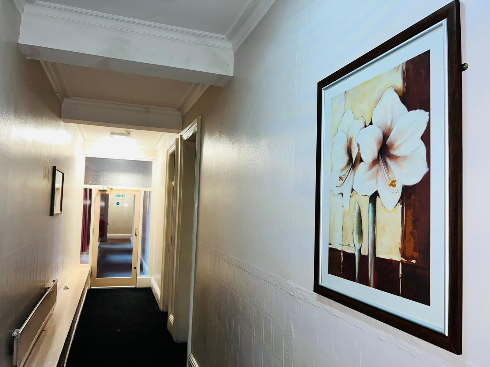 A hallway with a framed floral artwork on the wall, a door leading to a room, and a handrail on the left.