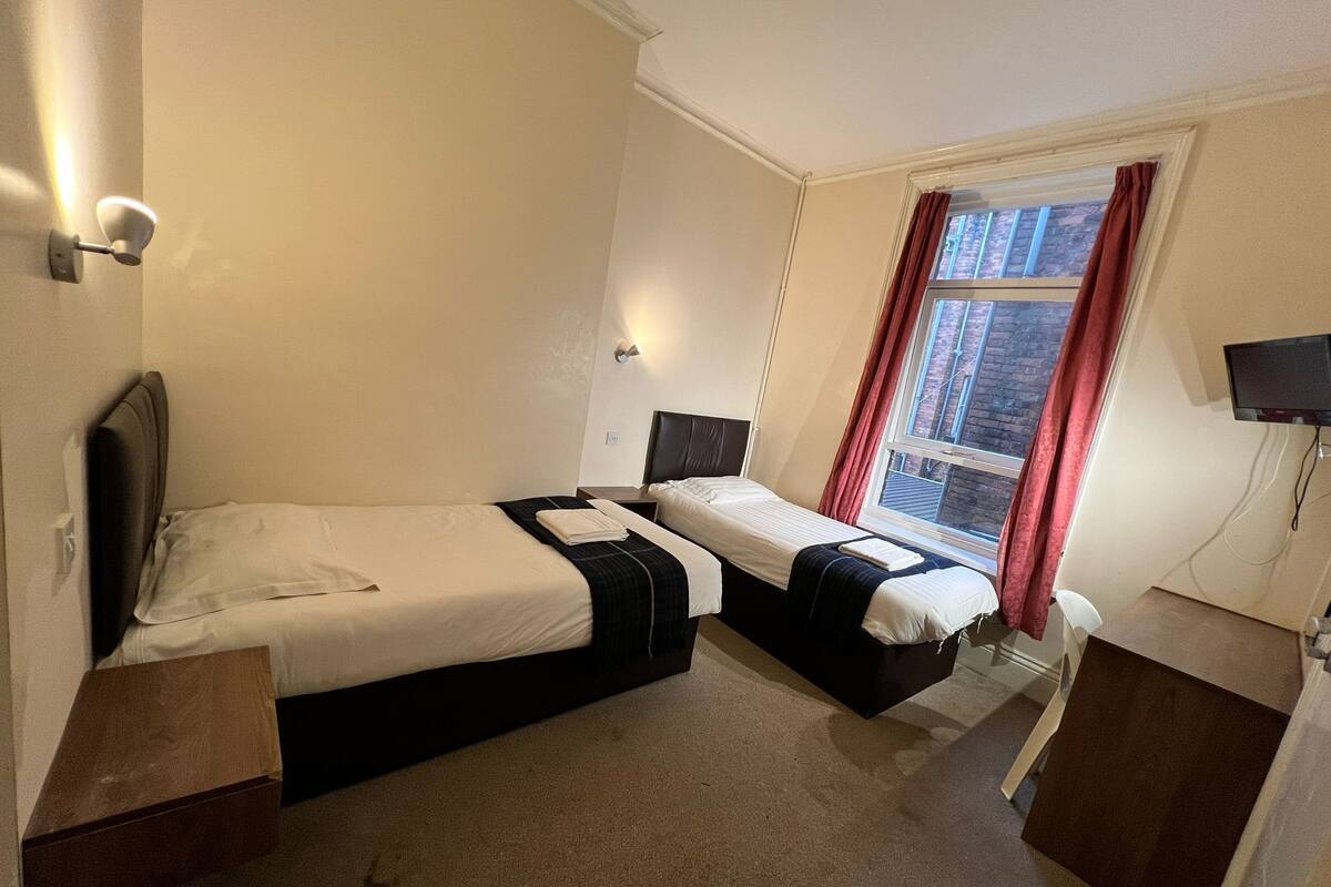 A hotel room with two beds, a window with red curtains, a flat-screen TV, and a small desk.