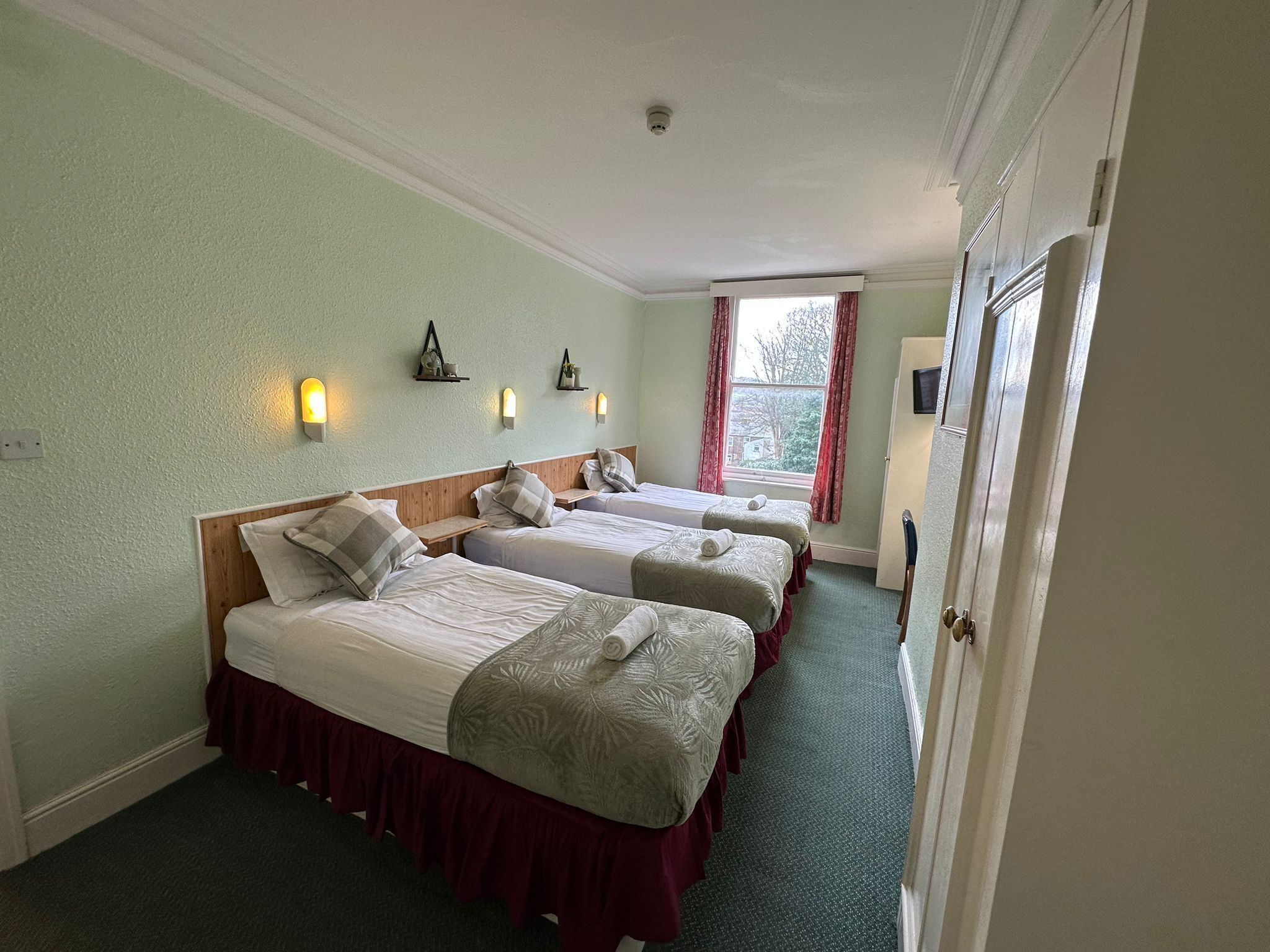 A hotel room with two beds, a window with curtains, wall-mounted lights, and a door.