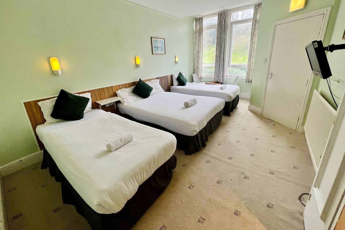 A hotel room with three single beds, a television, a window with curtains, and a radiator.