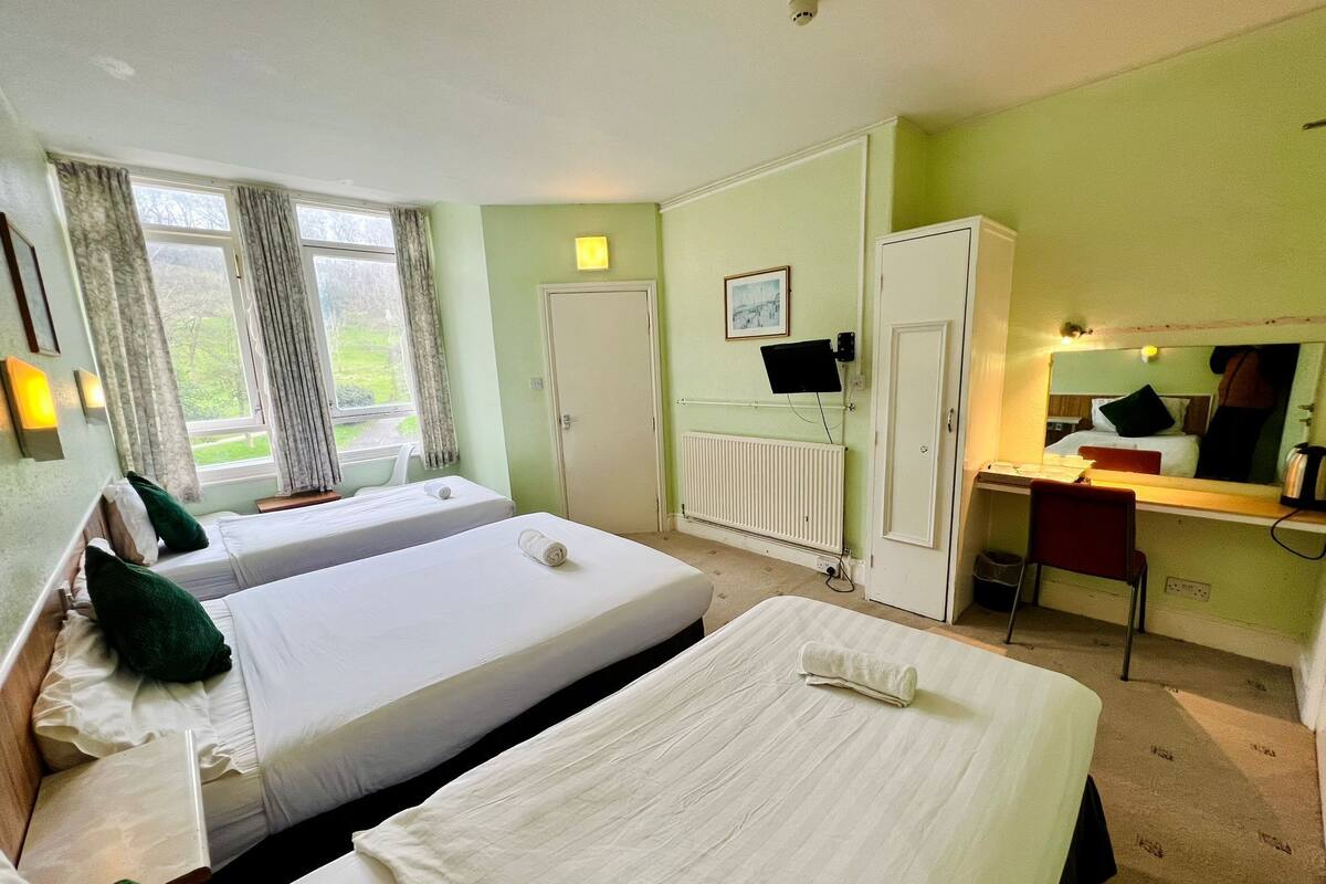A hotel room with two beds, a desk, a chair, a radiator, a television, and a window with curtains.